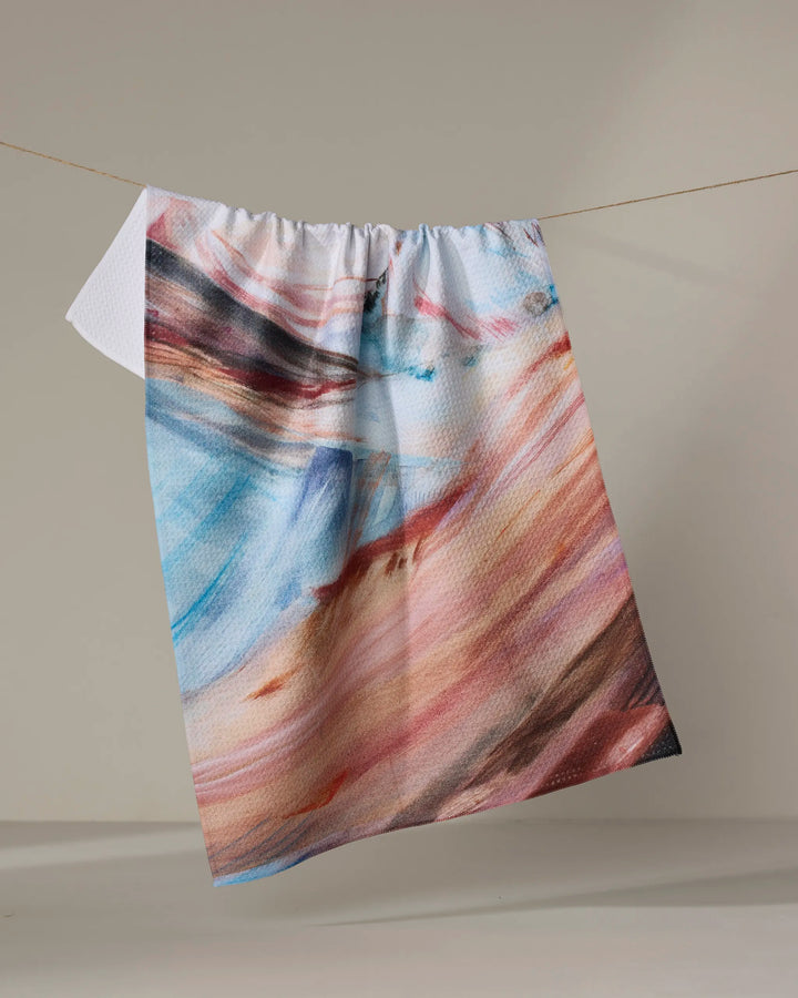 The "Golden Hour in the Rockies" tea towel by Kelly Peters features an abstract, colorful pattern in blue, pink, orange, and brown. Made from recycled materials for a sustainable touch, it hangs on a clothesline against a plain light background.