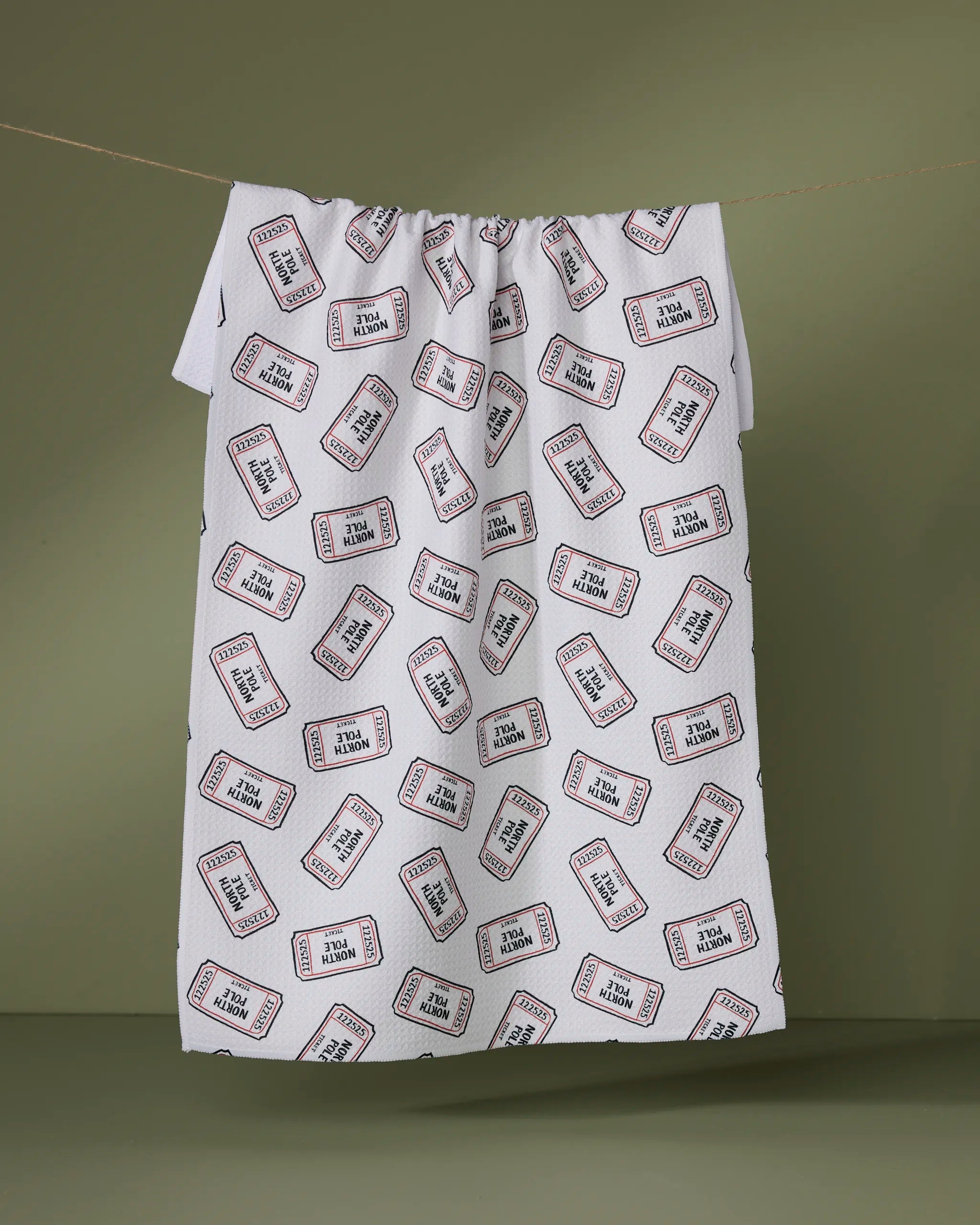 A white ultra-absorbent towel by Geometry, called Golden Pass, features repeating red and black "Admit One Woman" labels and hangs on a clothesline with an olive-green background.