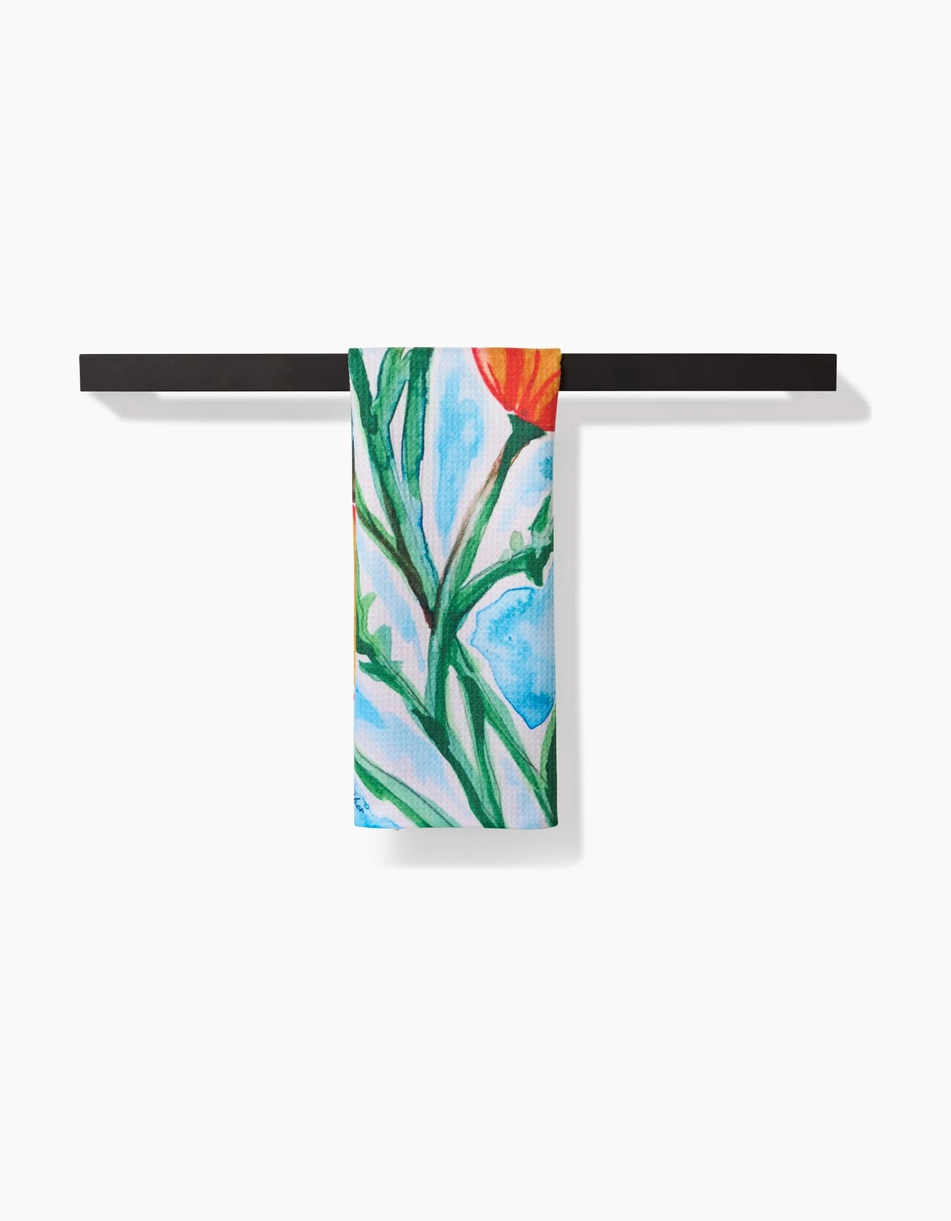 The Golden Poppy Flowers Tea Towel by Skye Walker hangs on a sleek, black bar, showcasing a vibrant floral design. Made from Geoweave™ fabric with post-consumer recycled materials, it features green stems, orange petals, and blue accents.