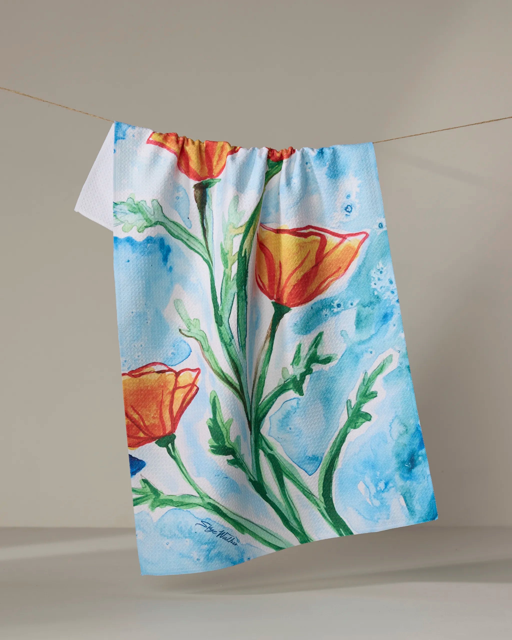 The Skye Walker "Golden Poppy Flowers" tea towel features orange poppies and green stems in a watercolor style on a blue background, hanging on a clothesline.