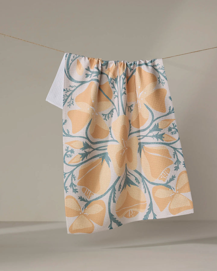 The "Golden Poppy" tea towel by Julianne Haness, featuring yellow and teal floral patterns and made from recycled materials, hangs over a clothesline against a neutral background.