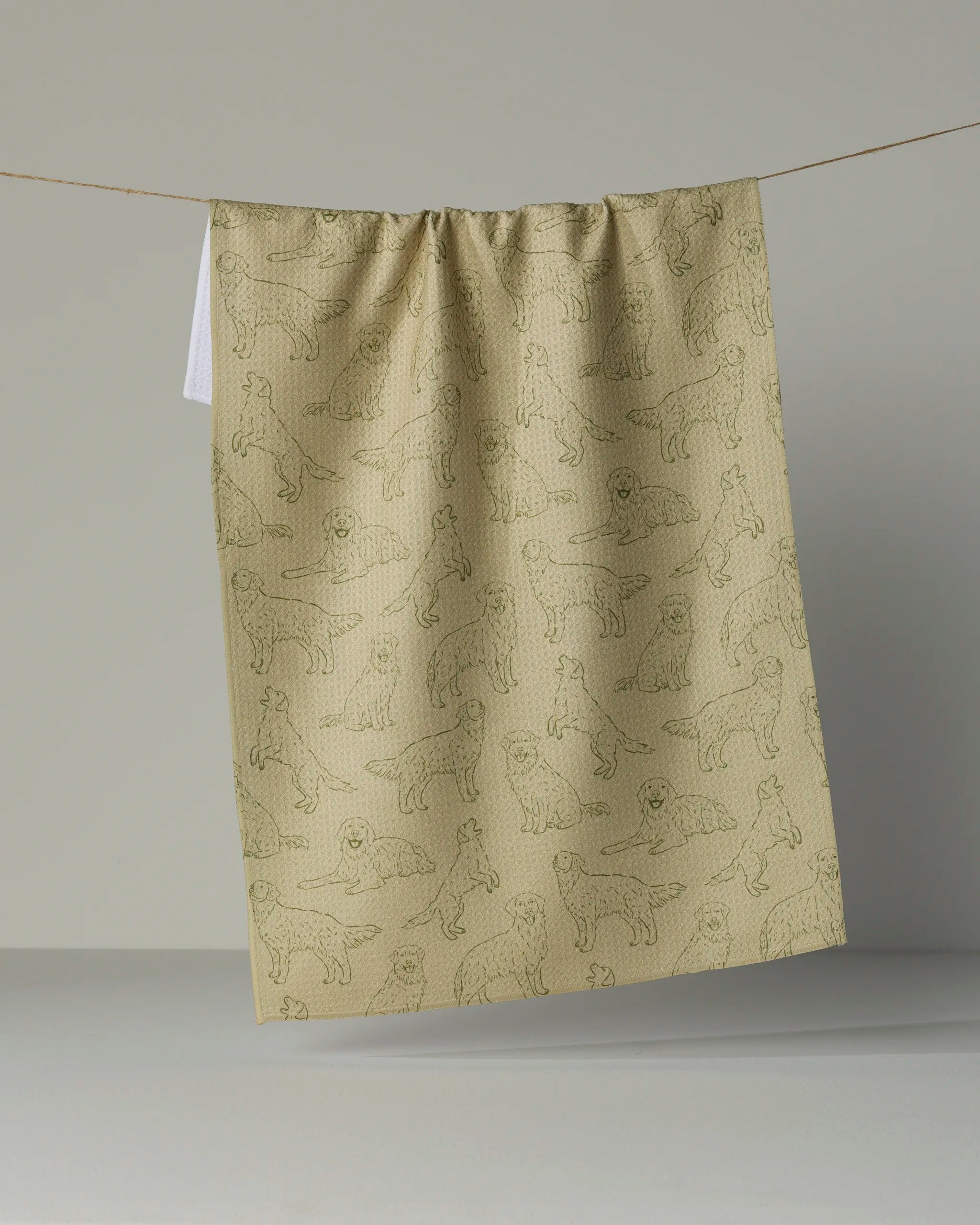 A “Golden Pups” tea towel by Geometry, crafted from recycled materials and adorned with line drawings of various dog breeds in beige, hangs on a string against a plain background.