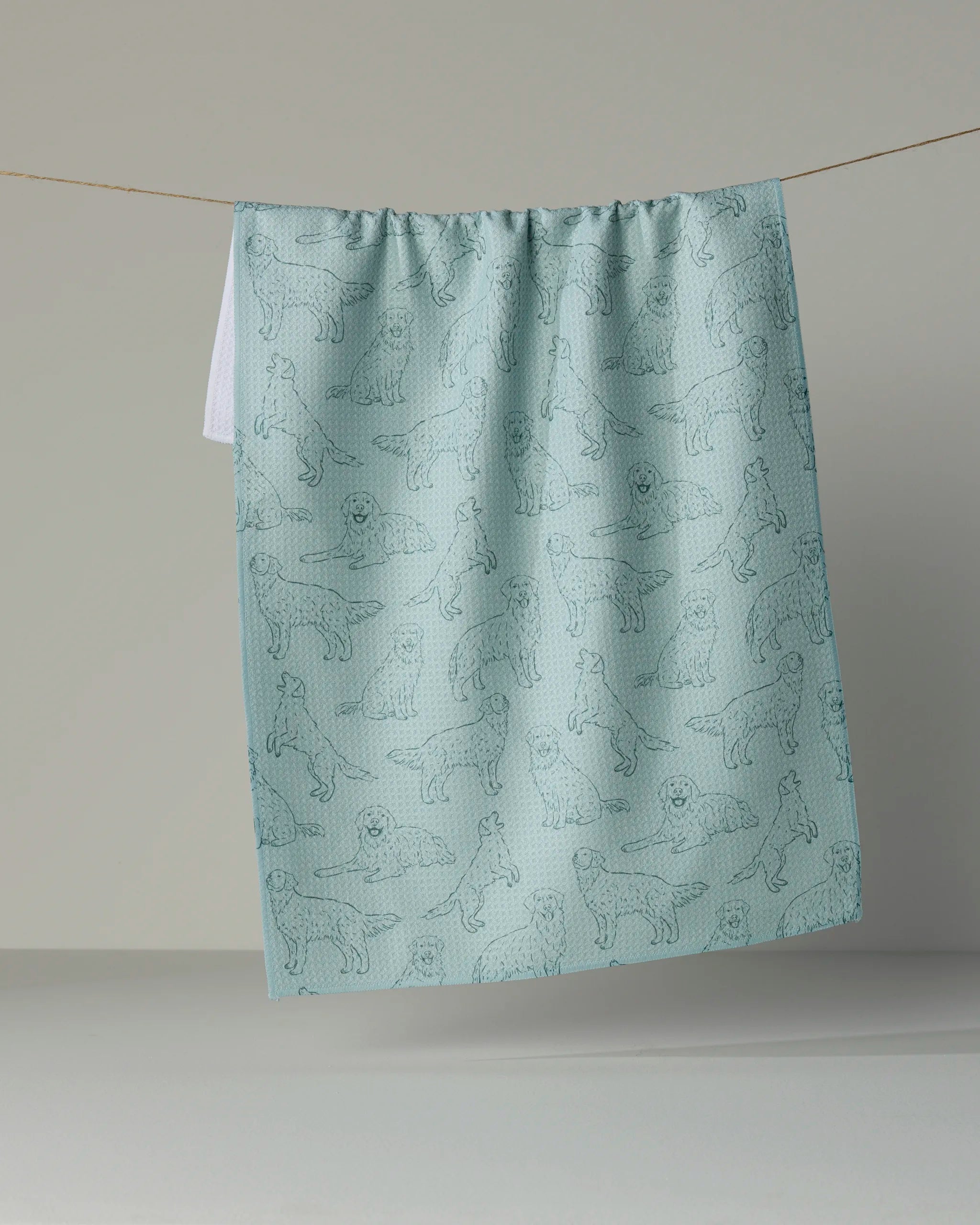 The “Golden Pups” kitchen towel by Geometry, crafted from recycled materials in a light blue hue, showcases an outline pattern of various dogs and hangs on a clothesline against a plain neutral background.