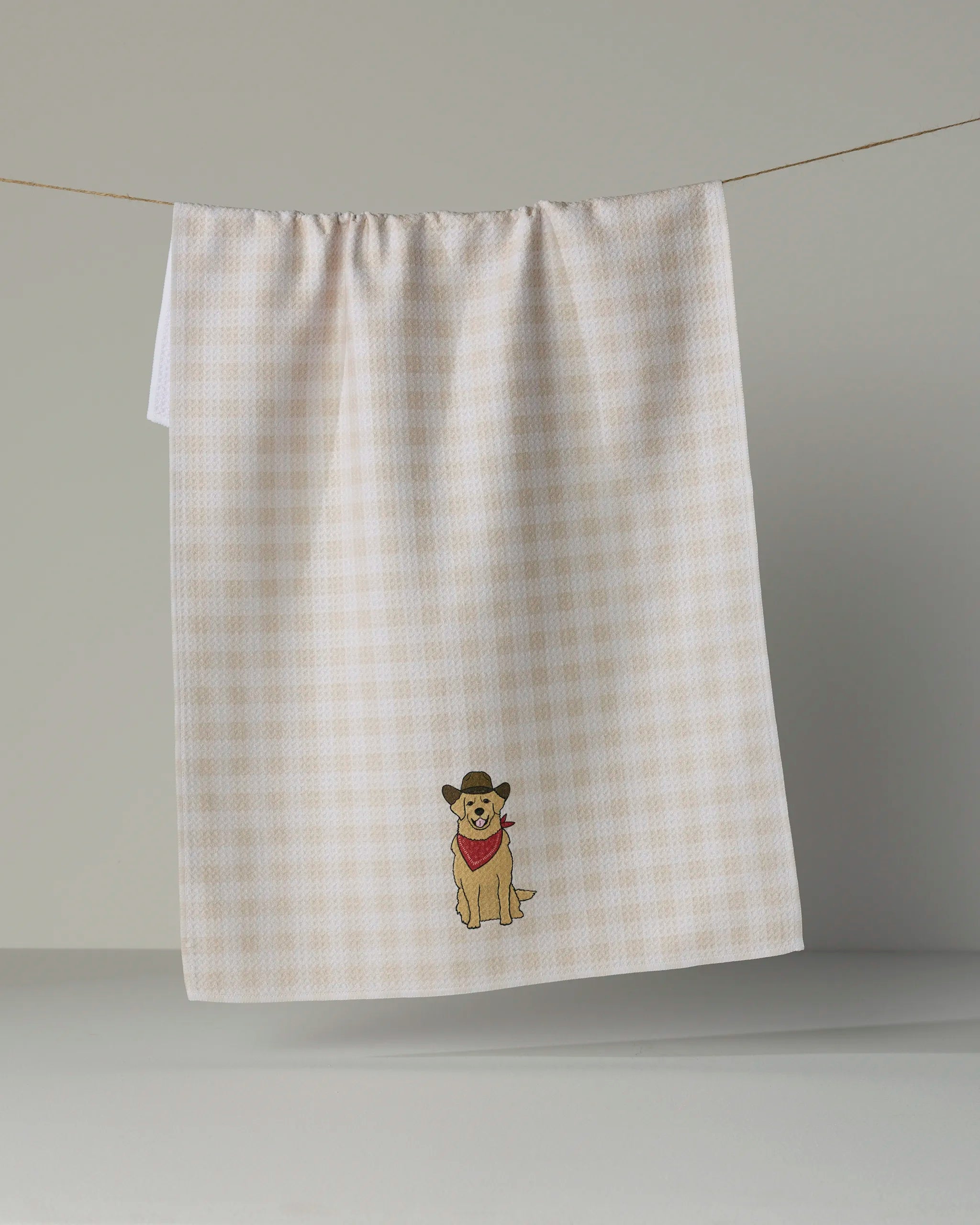 A beige and white checkered tea towel from Geometry, called “Golden Western,” hangs on a line. The ultra-absorbent towel features an embroidered design near the bottom center of a dog wearing a cowboy hat and red bandana.
