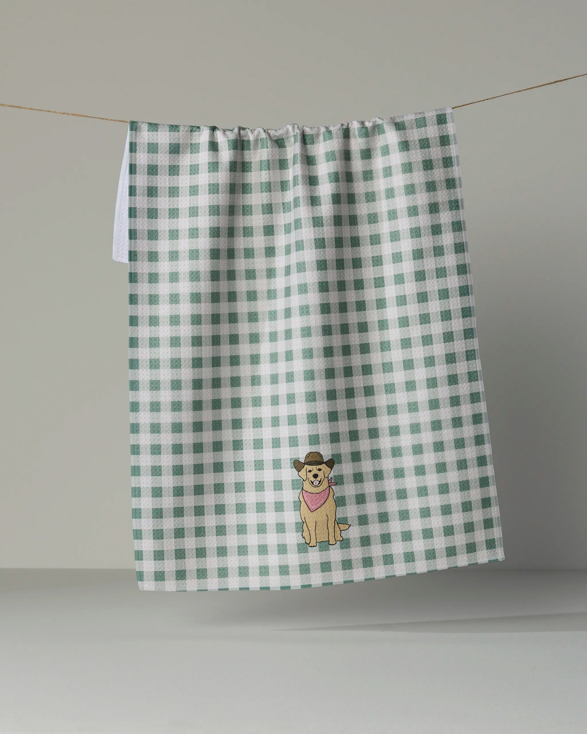 A tea towel from Geometry, the Golden Western, hangs on a line, showcasing a green and white checkered pattern. Ultra-absorbent and practical, it features an embroidered dog wearing a pink scarf near the bottom center, blending charming style with everyday function.
