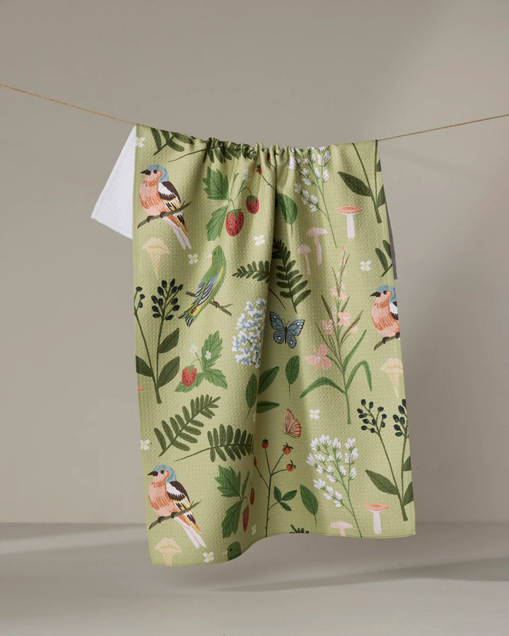 The "Good Green Earth" recycled kitchen towel by Janna Sue Design, with a botanical and bird print of flowers, leaves, butterflies, and finches, hangs on a string against a neutral background.