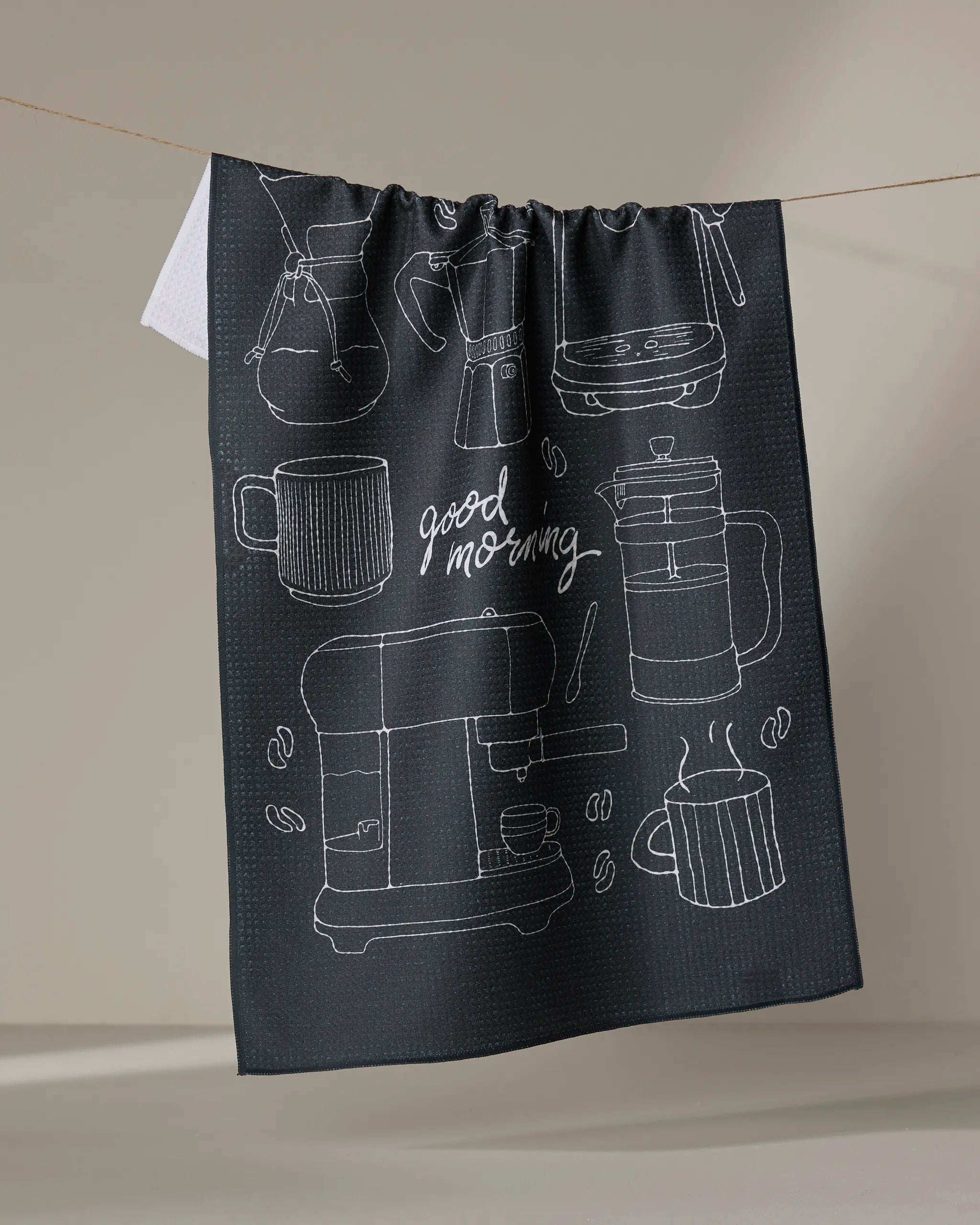 A dark recycled Rosa Kichline "Good Morning Coffee" kitchen towel hangs on a line, with white sketches of coffee makers, mugs, beans, and "good morning" in cursive.