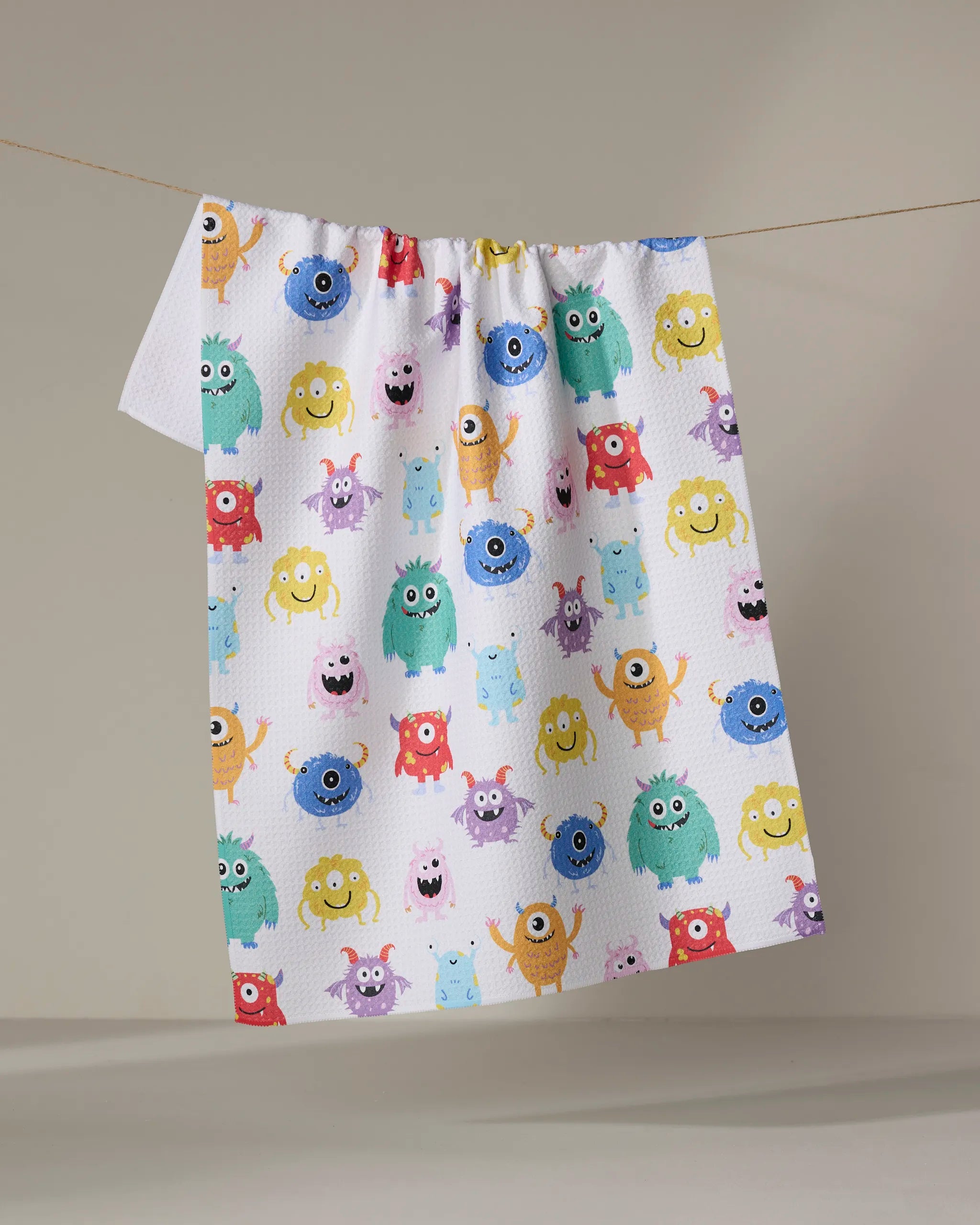 The Vaishali Design Studio "Goofy Critters" tea towel, ultra-absorbent and white, features colorful cartoon monsters with playful expressions. It hangs on a clothesline against a plain beige background.