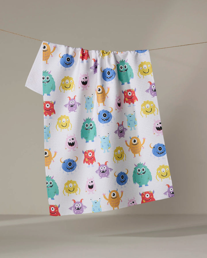 The Vaishali Design Studio "Goofy Critters" tea towel, ultra-absorbent and white, features colorful cartoon monsters with playful expressions. It hangs on a clothesline against a plain beige background.