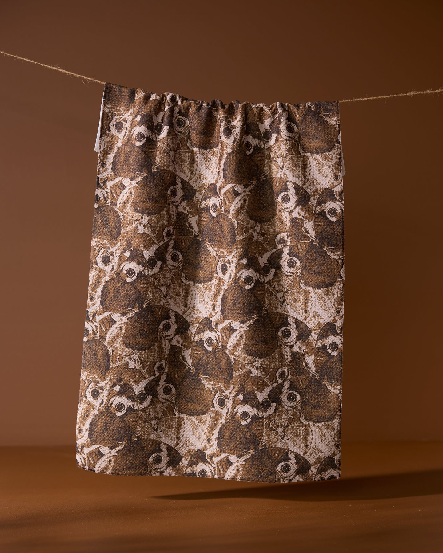 The Geometry Gossamer Flight tea towel, made from soft Geoweave fabric, features an illustrated pattern of owl faces and hangs on a string against a solid brown background.