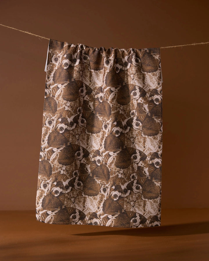 The Geometry Gossamer Flight tea towel, made from soft Geoweave fabric, features an illustrated pattern of owl faces and hangs on a string against a solid brown background.