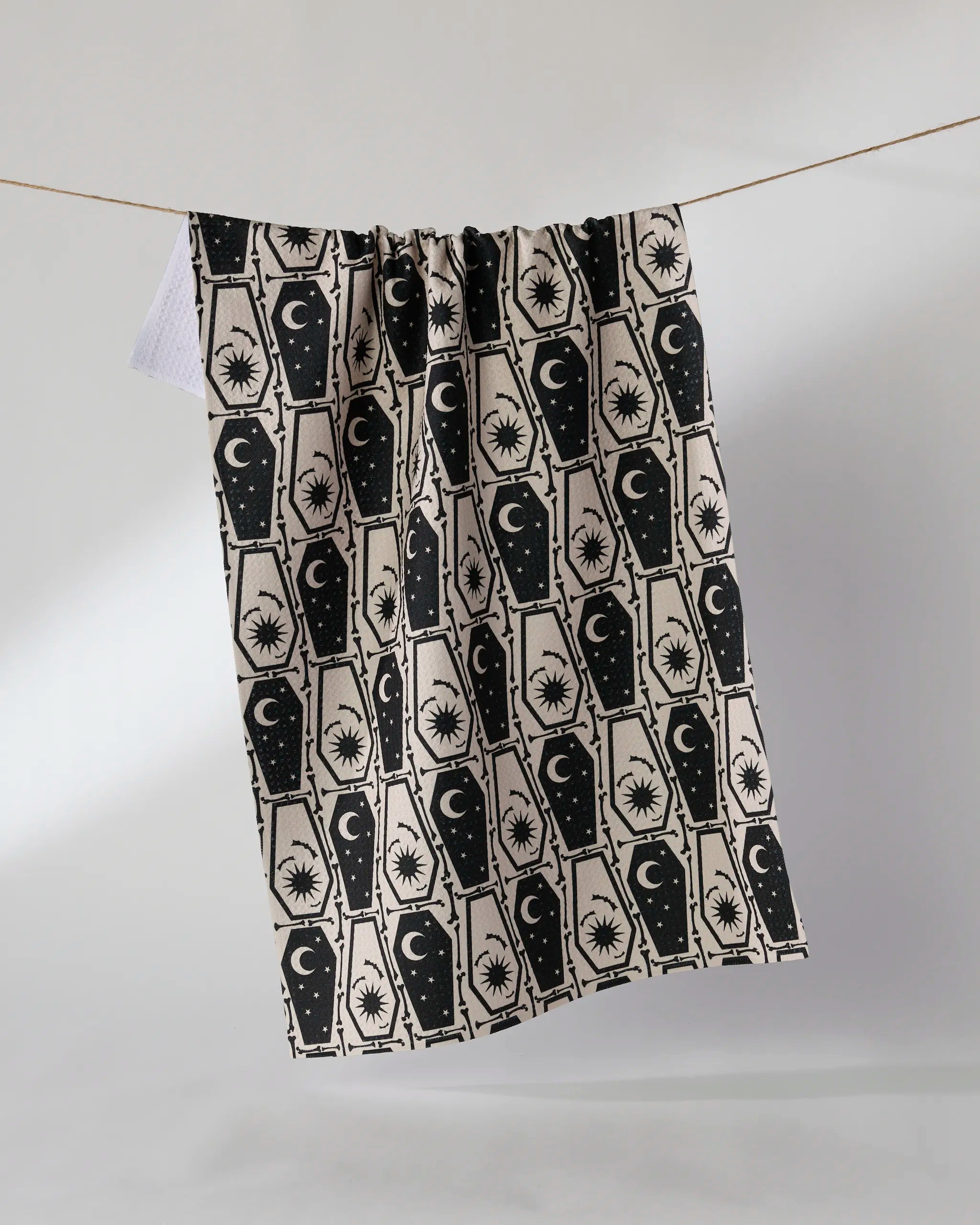 The Gothic Slumber kitchen towel by Geometry, featuring a repeated black pattern of coffins, crescent moons, stars, and suns on beige recycled fabric, hangs on a string against a plain light background.