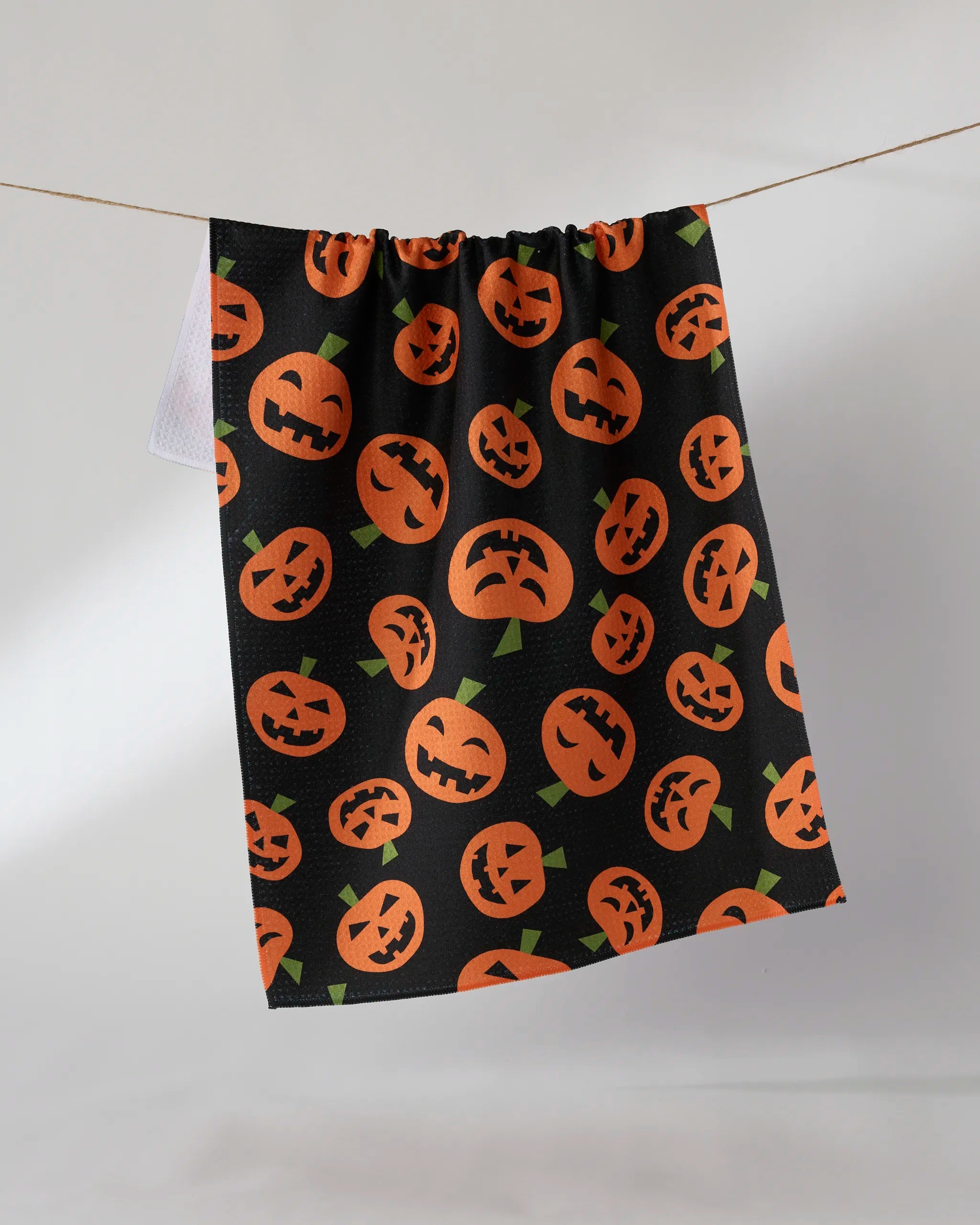 The Geometry "Gourd Gang" is a black tea towel made from recycled materials, decorated with orange jack-o-lantern faces in various expressions, displayed against a light background.