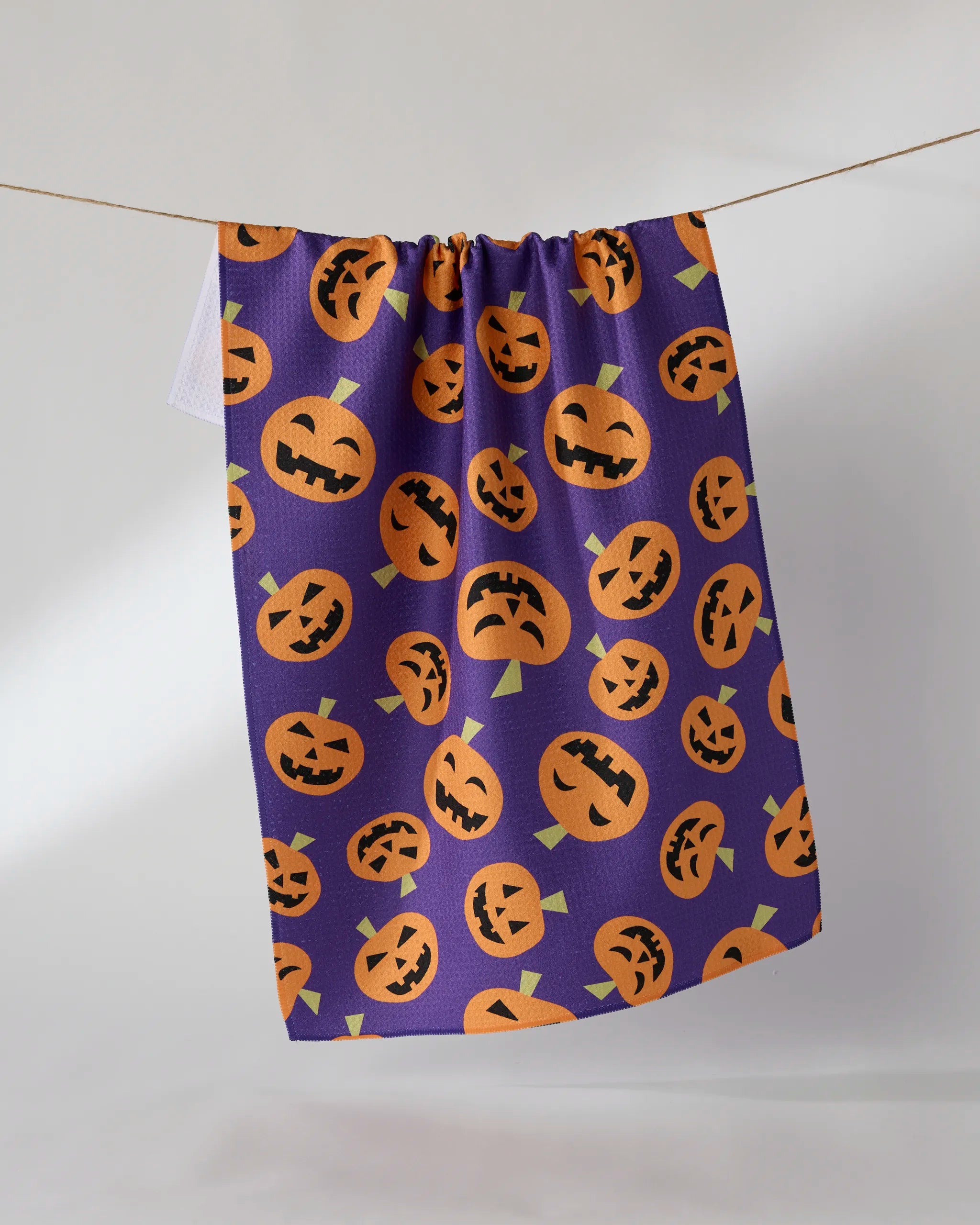 The Geometry Gourd Gang tea towel hangs on a clothesline, showcasing a purple background with orange jack-o-lanterns in various expressions, and is crafted from recycled materials.