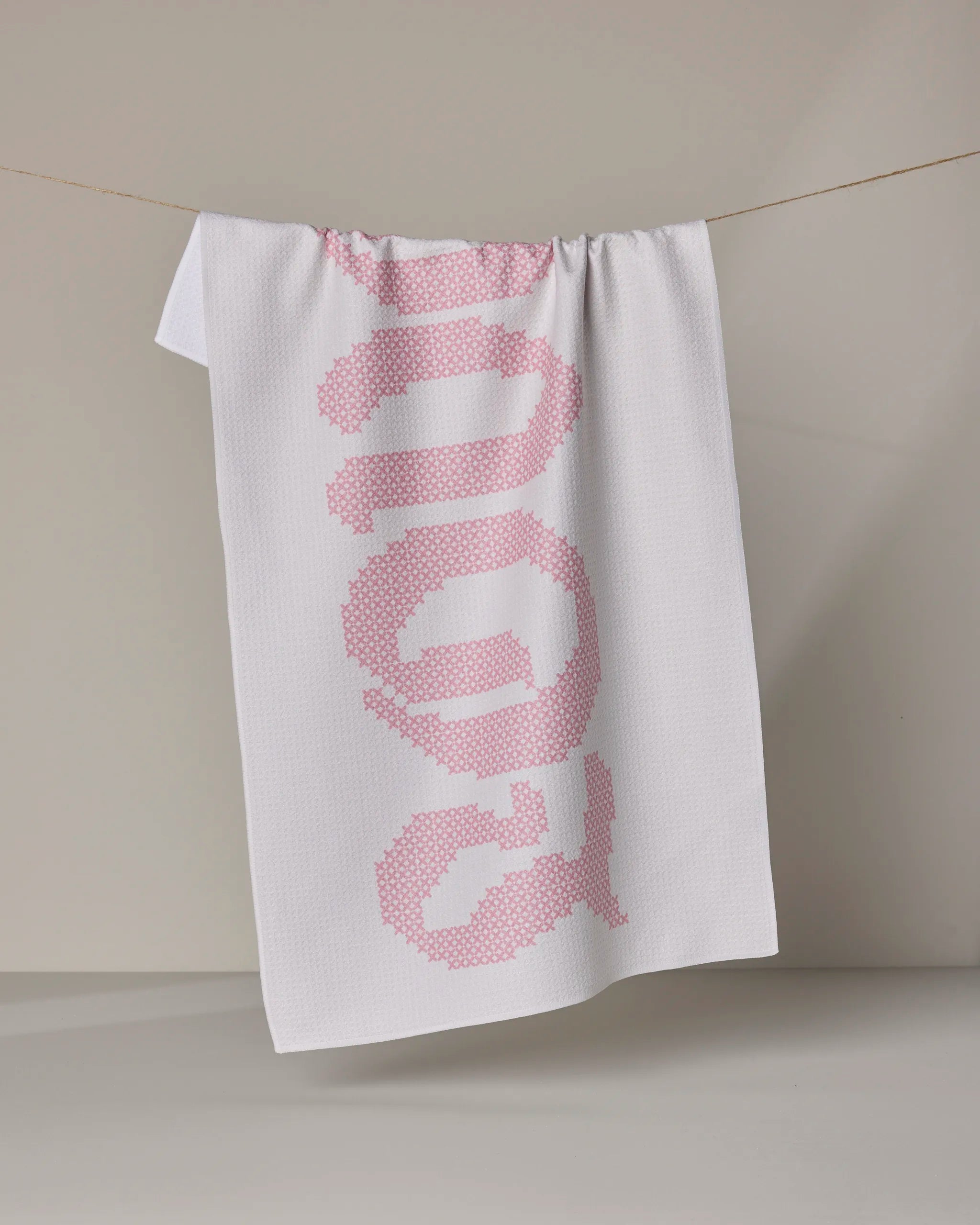 The Grand Love White tea towel by Geometry, made from recycled materials, hangs on a clothesline with “nope” stitched vertically in bold pink letters against its neutral backdrop, emphasizing the playful design.