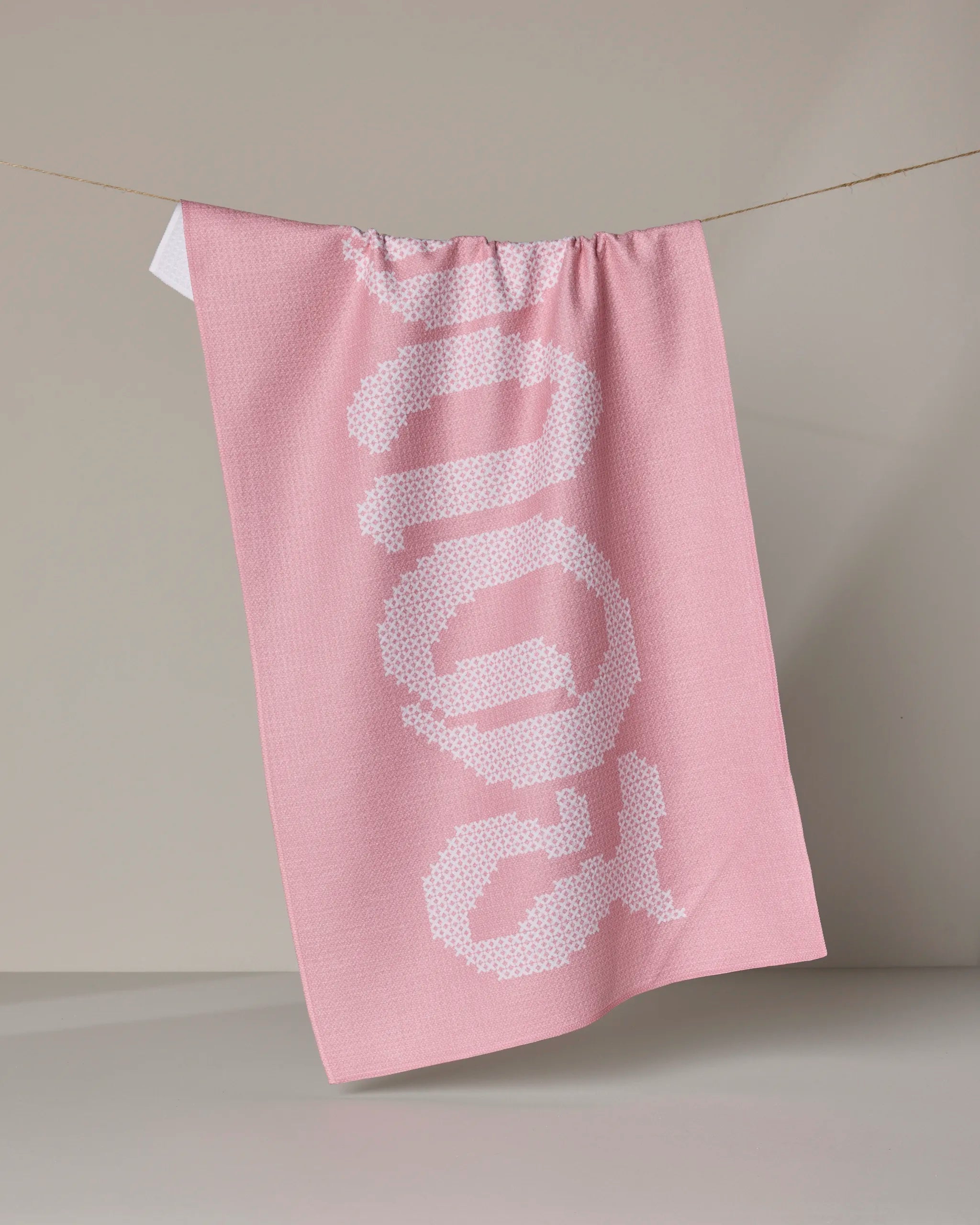 A "Grand Love" tea towel by Geometry, made from recycled materials, in pink with "DOG" in large white dotted letters, hangs on a clothesline against a neutral background.