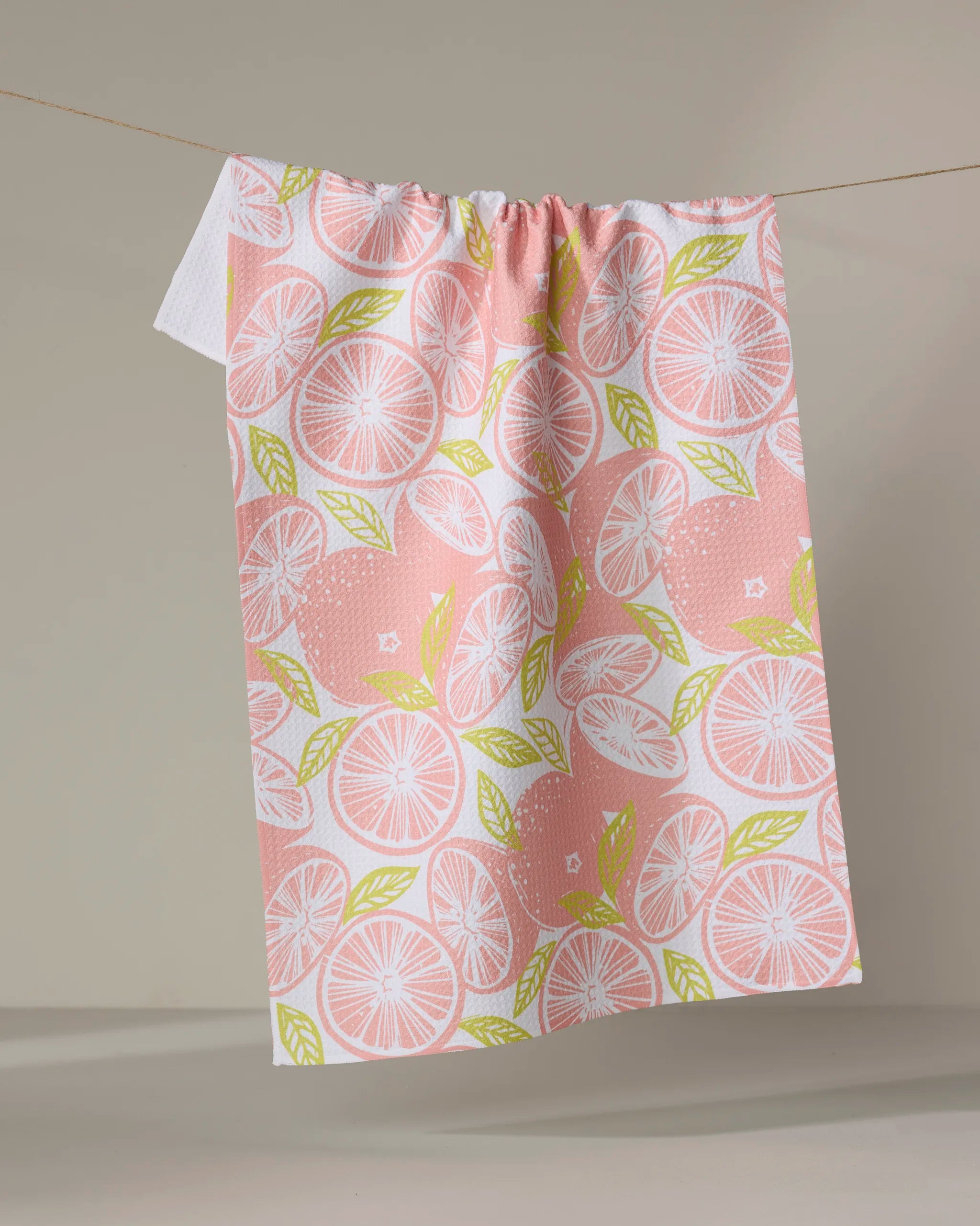 The "Grapefruits" kitchen towel by Elisabeth Wing, made from recycled materials and decorated with a pink and green citrus fruit pattern, hangs on a string against a plain beige background.