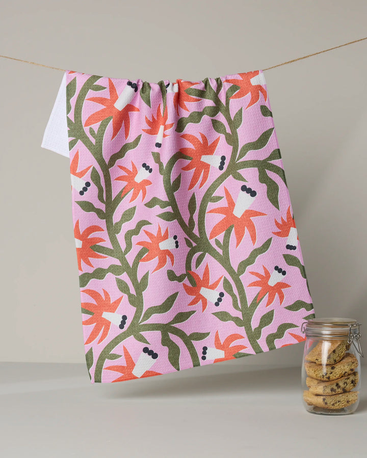 The Claire Iglesias "Graphic Flowers" tea towel, featuring a pink background with orange and green floral patterns and dark berries, made from recycled materials, hangs on a string beside a glass jar of cookies.