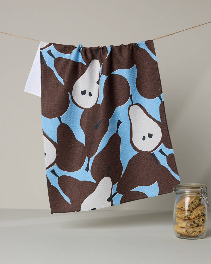 The Graphic Pears kitchen towel by Claire Iglesias, featuring a brown, blue, and white pear pattern, hangs on a string above a jar of cookies on a white surface against a neutral background.