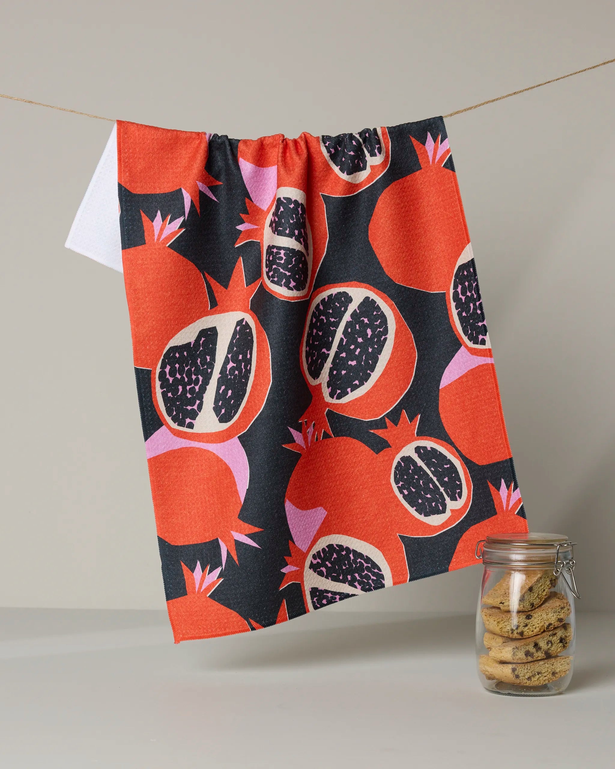 The Graphic Pomegranates tea towel by Claire Iglesias, featuring a bold red and black pomegranate pattern, hangs above a small glass jar of cookies against a neutral background.