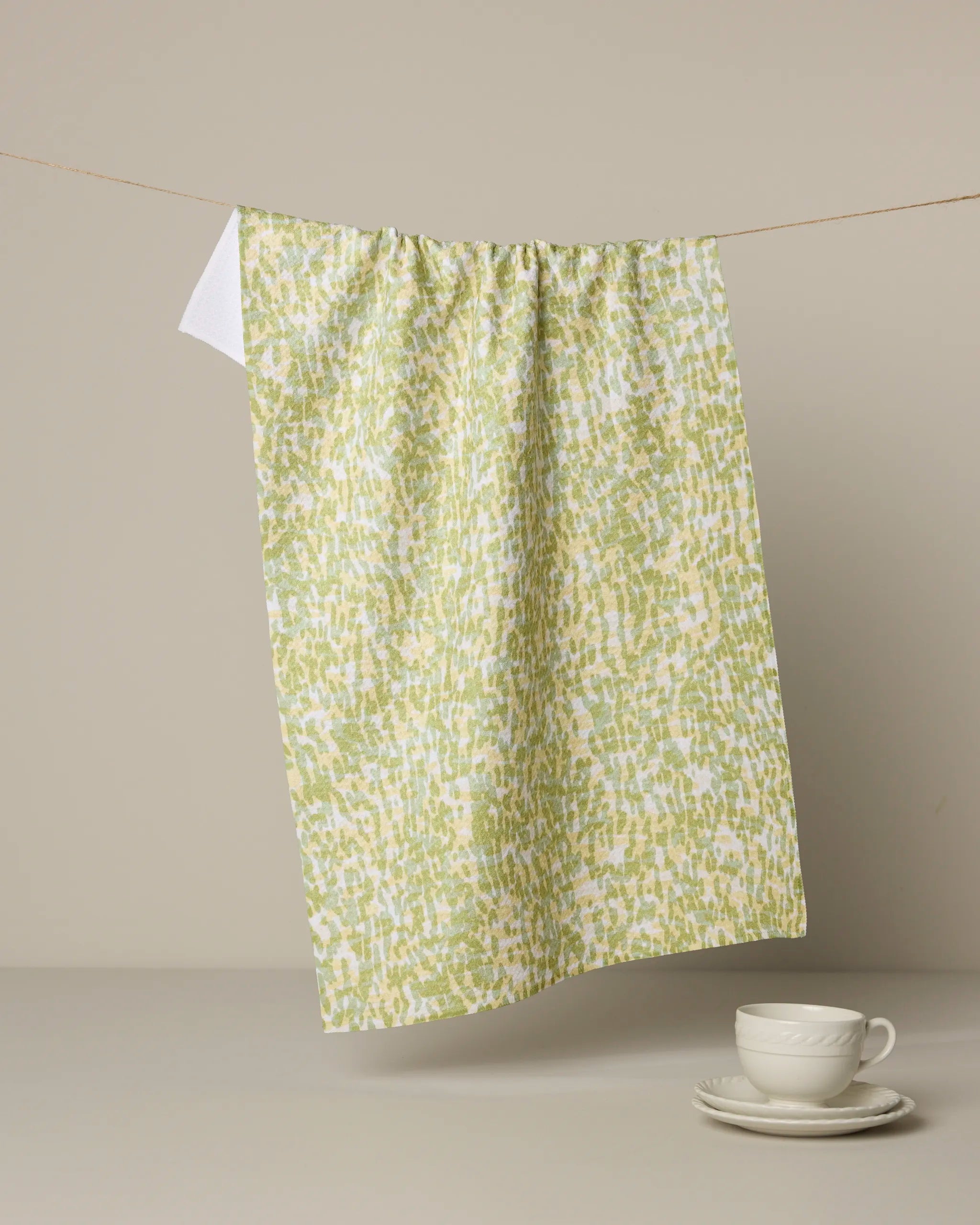 The Grassy Field tea towel by Geometry, featuring green and white dots, hangs from a string against a neutral background. Beneath it, a white teacup and saucer rest on a gray surface.