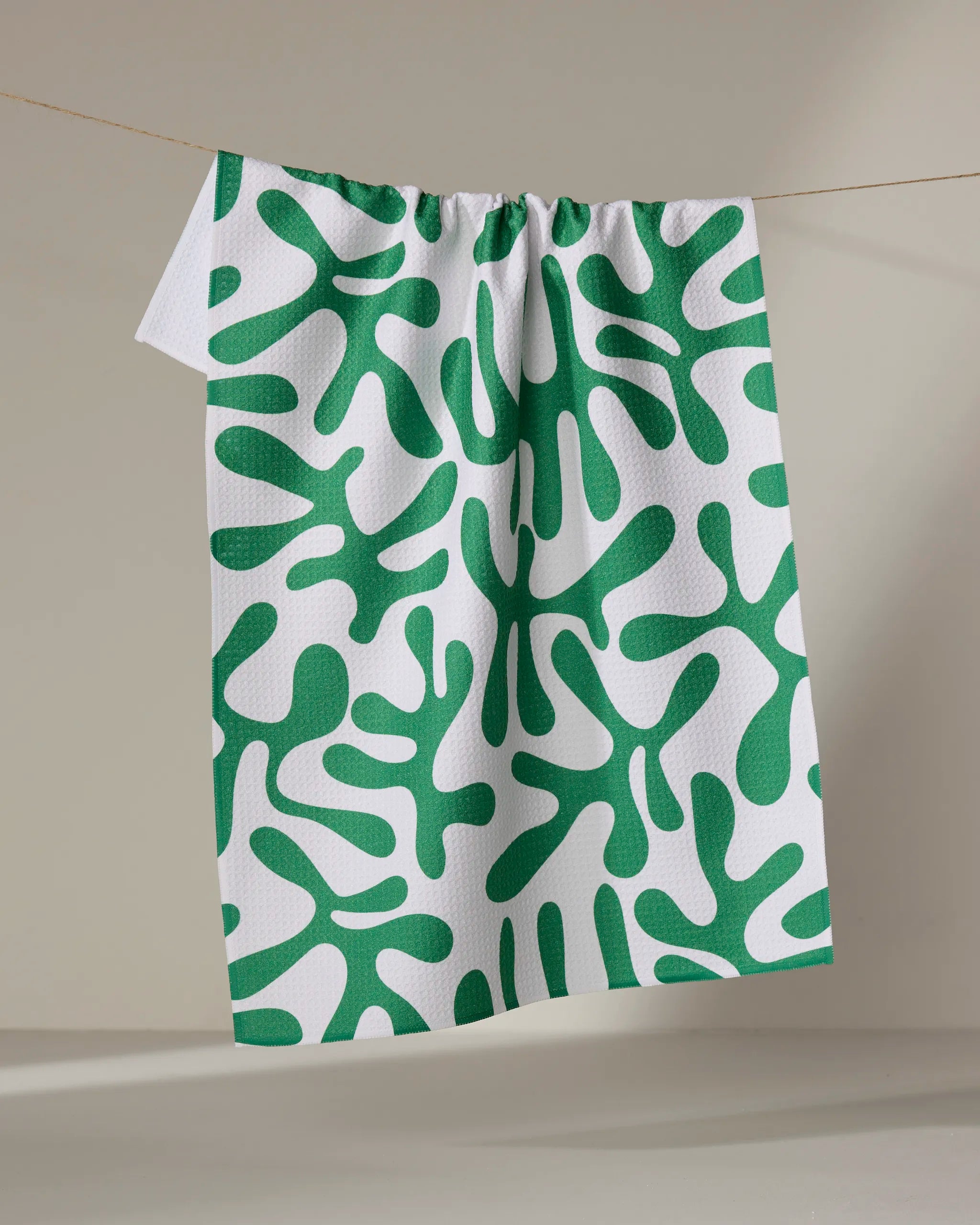 The Green Afternoon tea towel by Marina Solodka features an abstract green organic pattern and is made from recycled materials. It hangs on a thin string against a neutral background.