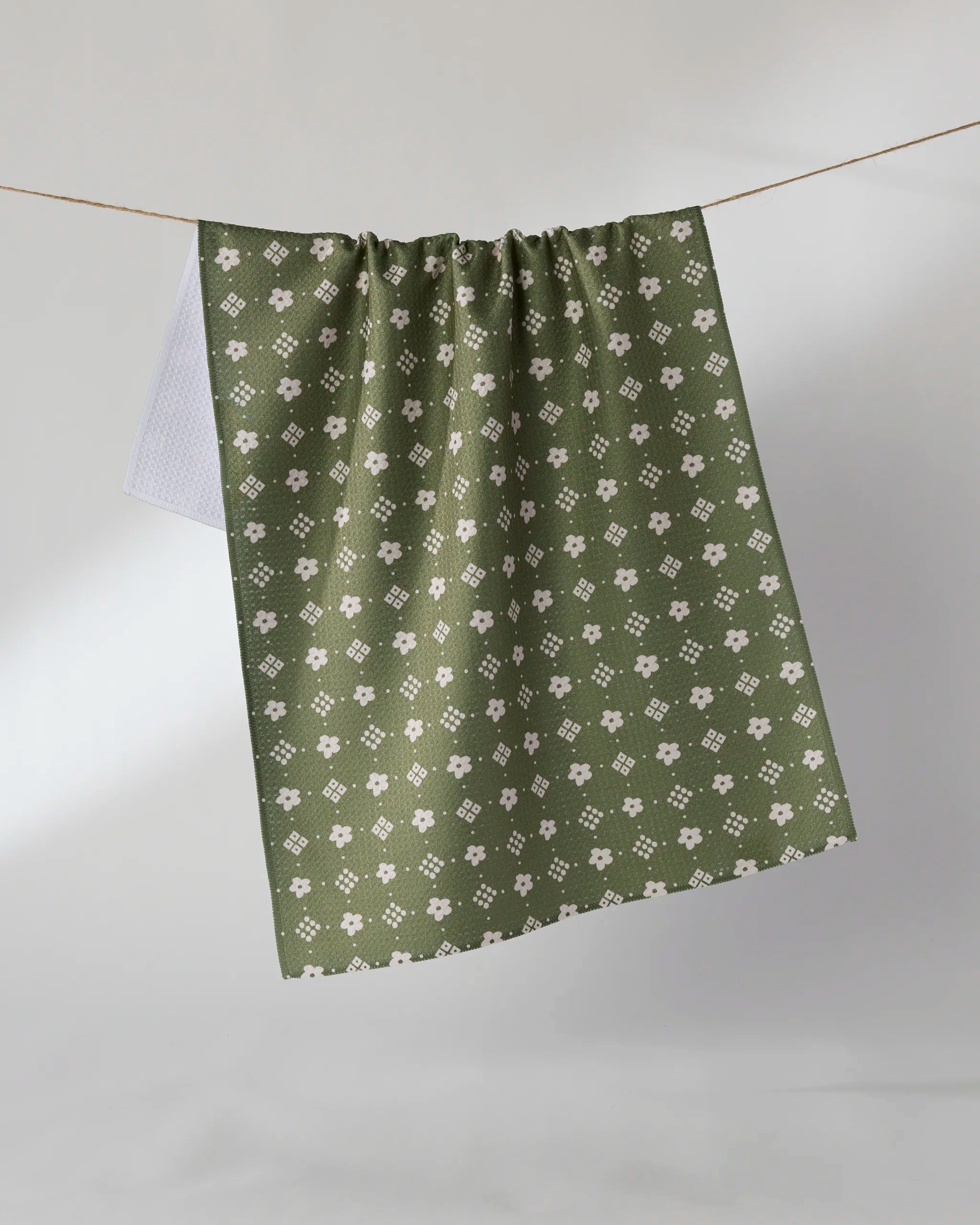 The Geometry Green Gables tea towel, featuring a white floral print and crafted from recycled materials, hangs on a clothesline against a light background.