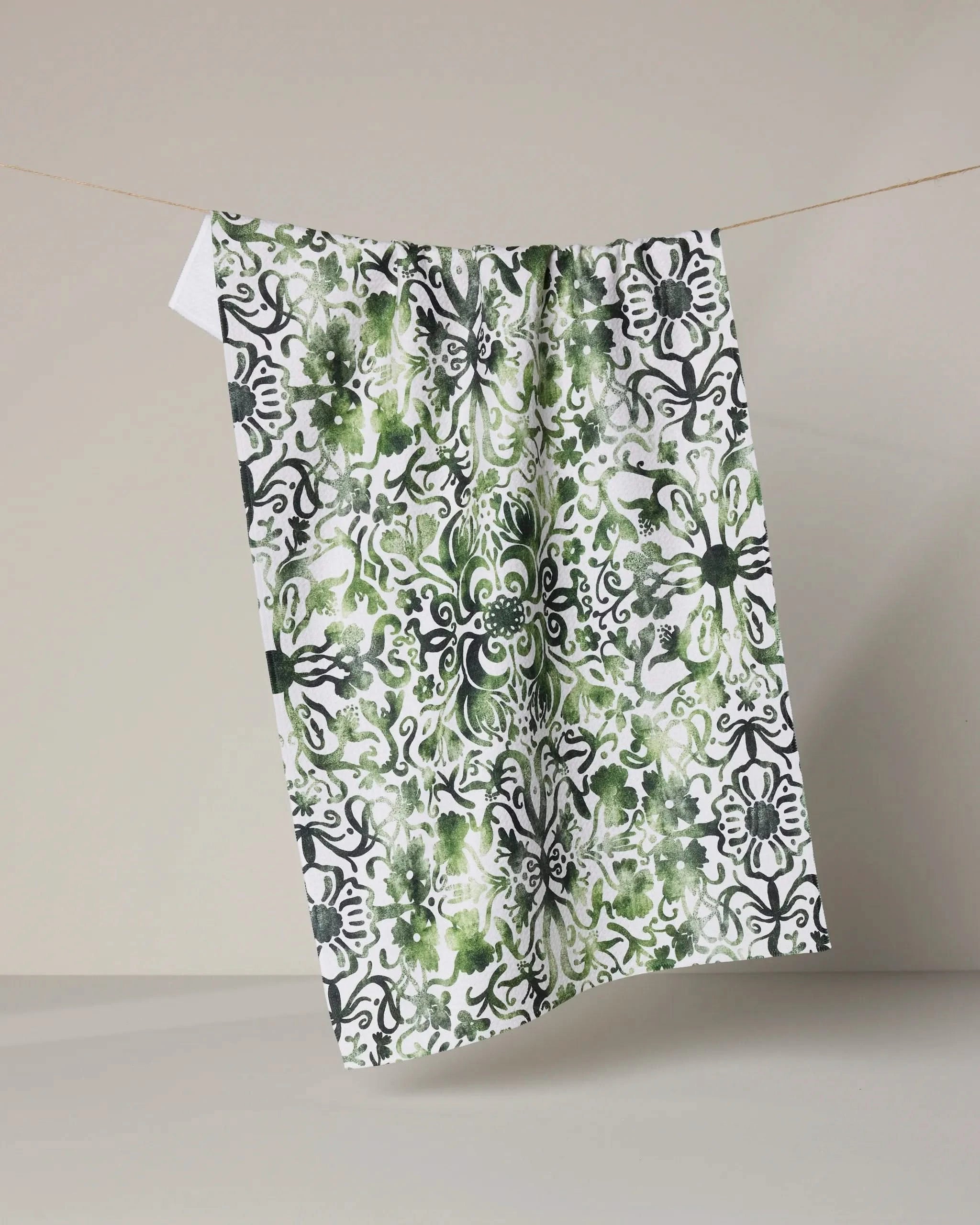 The Green Goddess tea towel by Geometry, featuring an ultra-absorbent design with a detailed black and green floral pattern, hangs from a thin line against a plain beige background.