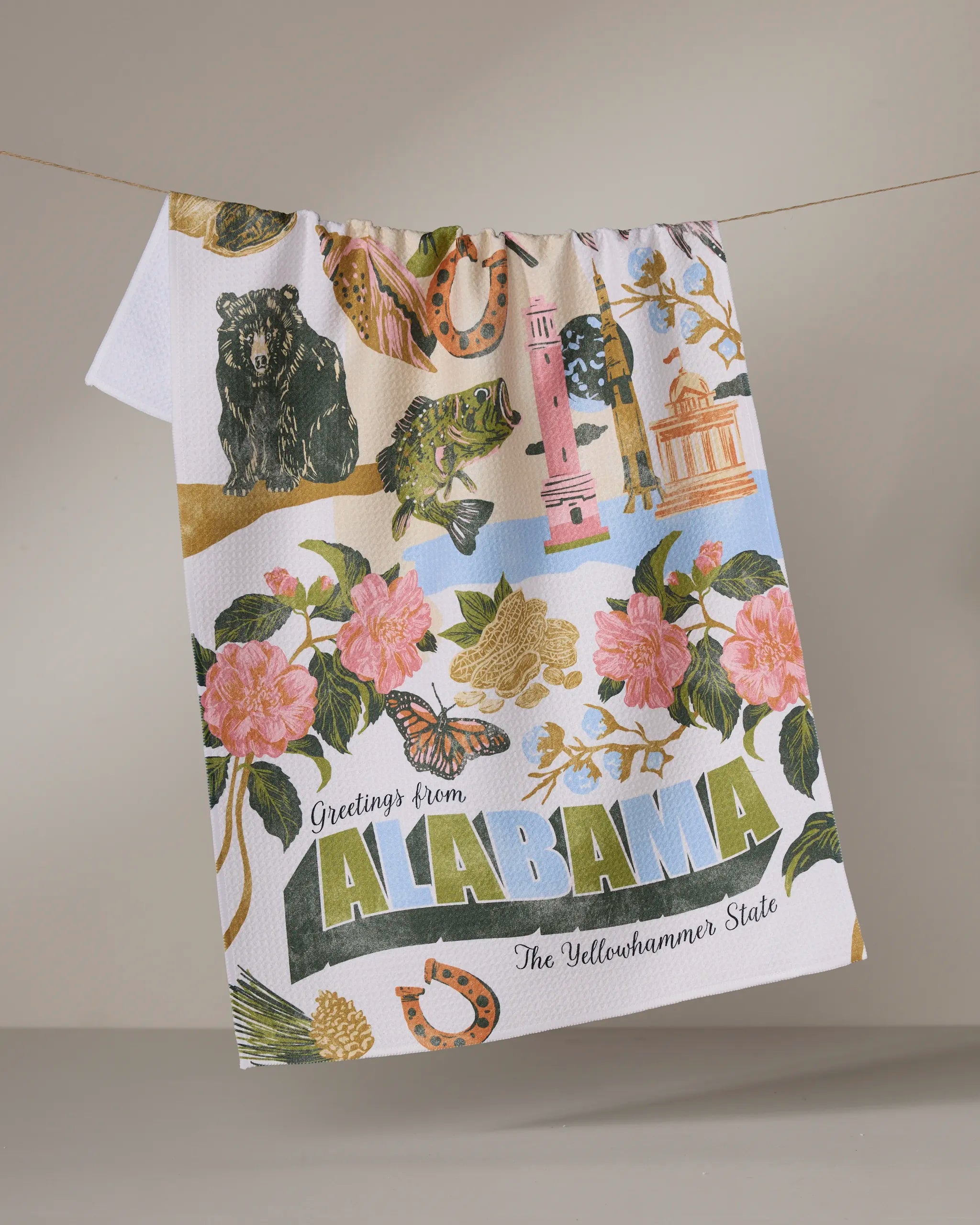 The Geometry "Greetings From Alabama" tea towel, ultra-absorbent and colorful, hangs on a line featuring Alabama symbols—flowers, animals, landmarks—and the text “Greetings from ALABAMA The Yellowhammer State.”.