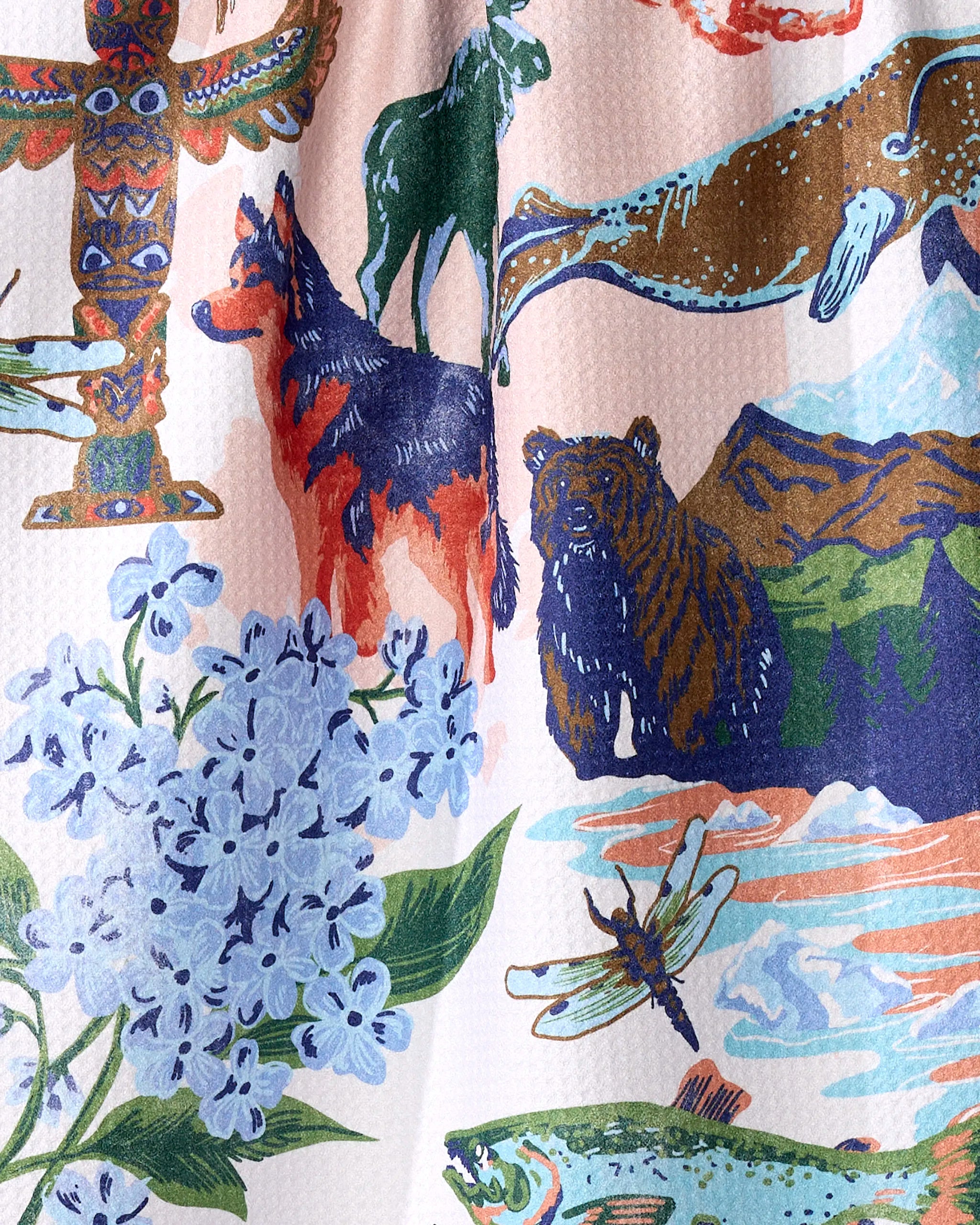 The Geometry "Greetings From Alaska" tea towel features totem pole, wolf, bear, whale, flowers, dragonfly, and mountain landscape illustrations on a light background—crafted from recycled materials for an eco-friendly touch.