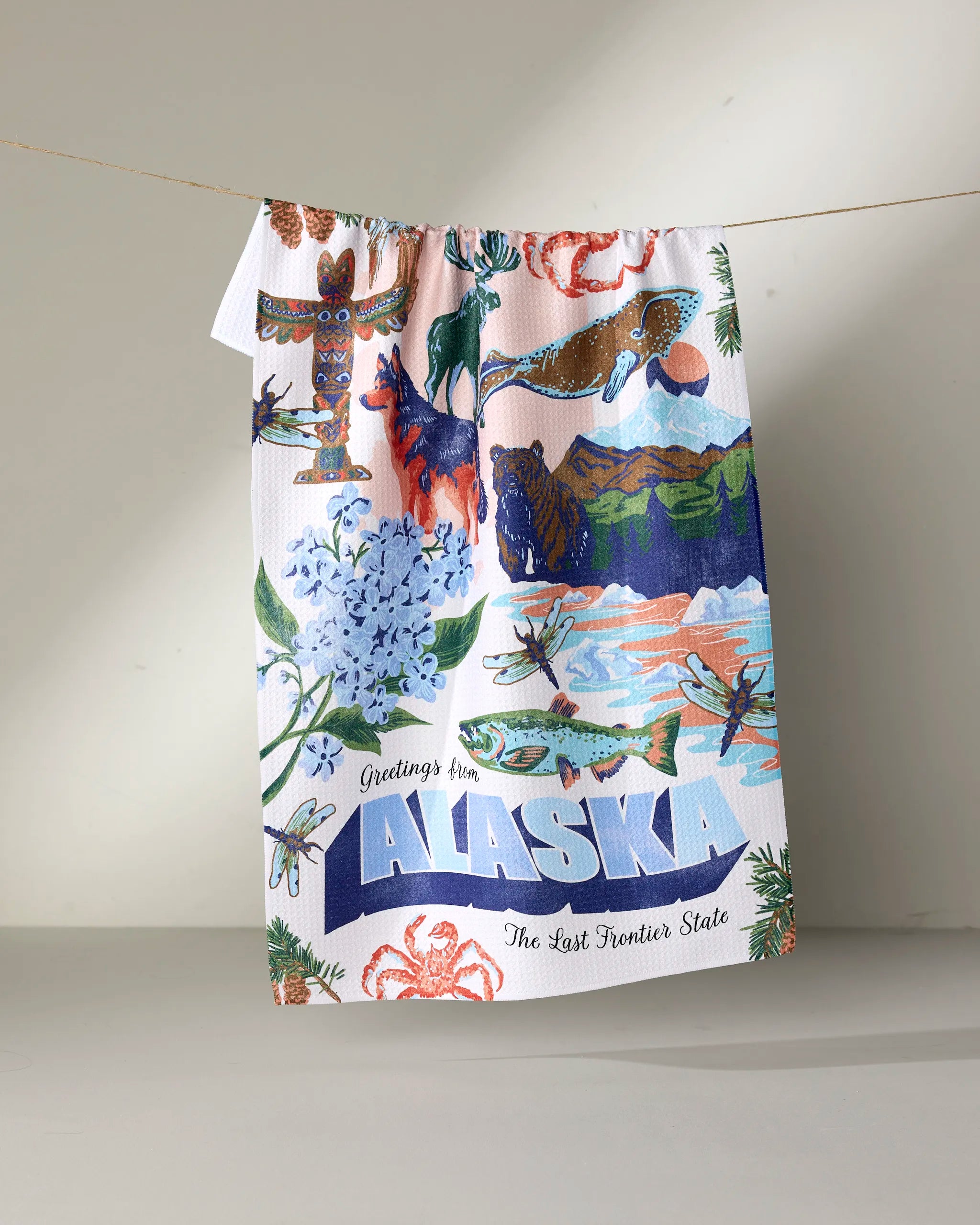 The Geometry "Greetings From Alaska" tea towel features colorful illustrations of Alaska wildlife, mountains, flowers, totem poles, and fish. Text reads "Greetings from ALASKA - The Last Frontier State." Made with recycled materials.