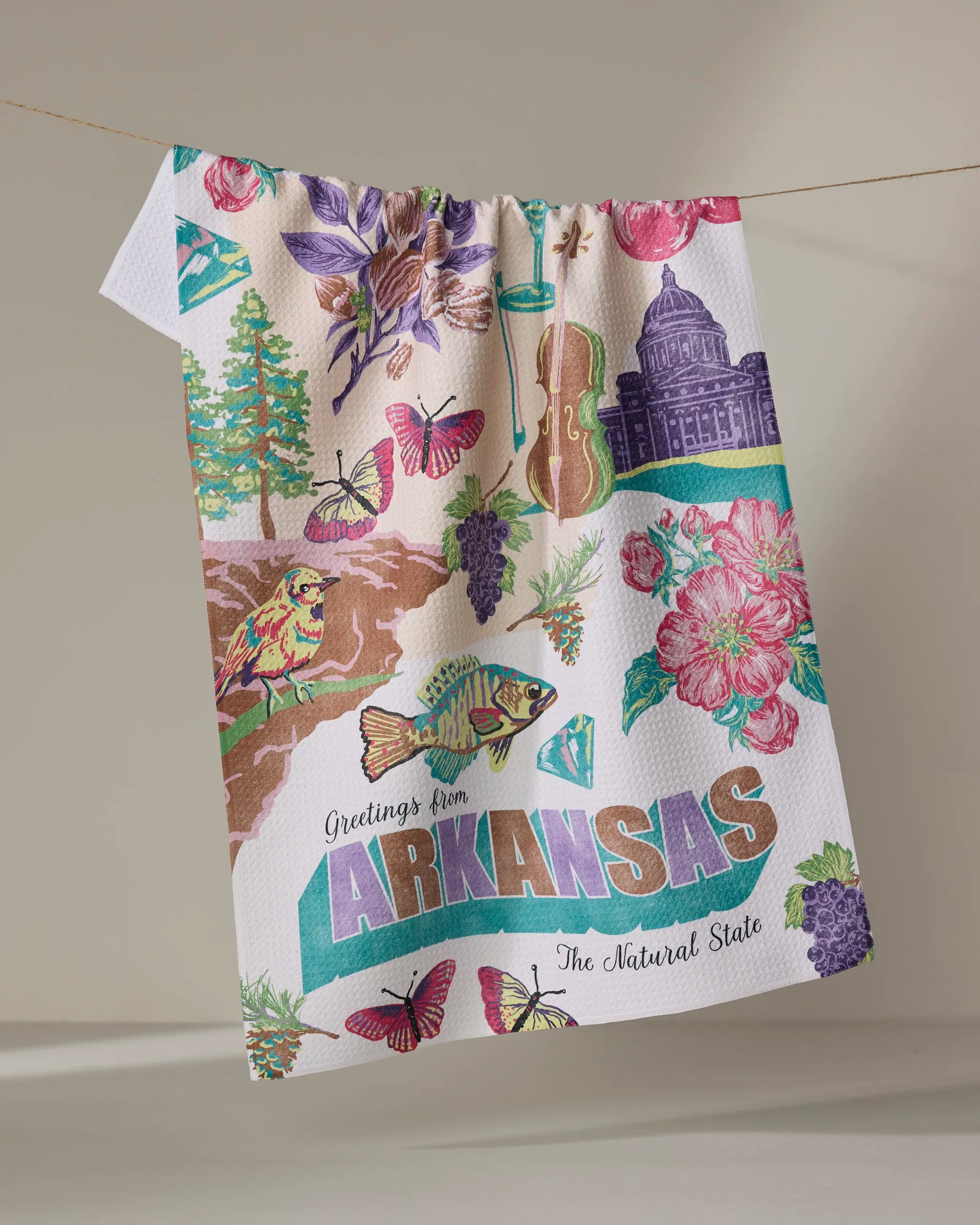 The Geometry "Greetings From Arkansas" tea towel, made from recycled materials, features Arkansas-themed illustrations—flowers, butterflies, fish, a diamond, trees, the state capitol, and “Greetings from Arkansas, The Natural State.”.