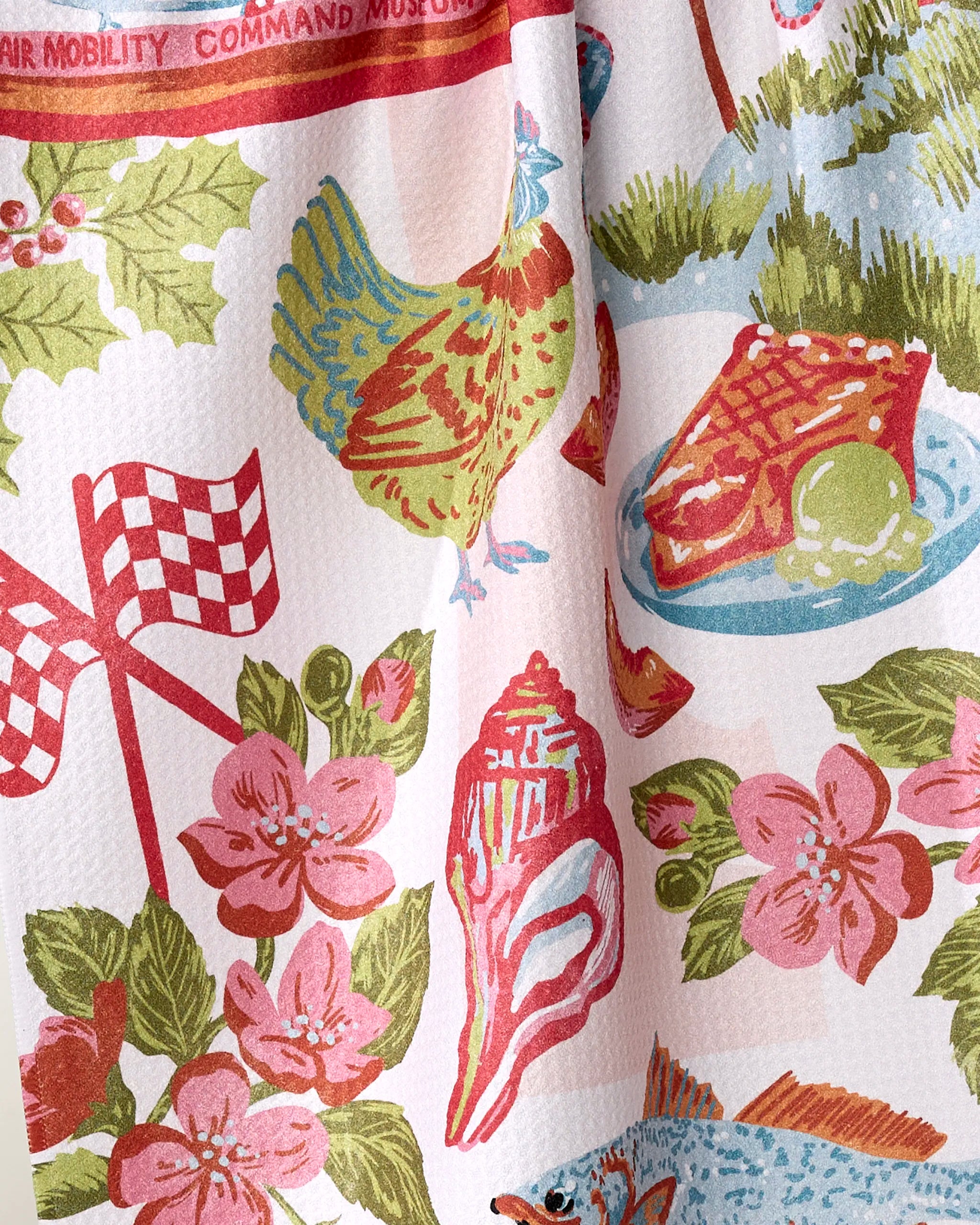 The “Greetings From Delaware” tea towel by Geometry is made from recycled materials and features vibrant retro-style illustrations of a chicken, steak, pink flowers, green leaves, checkered flags, and holly.