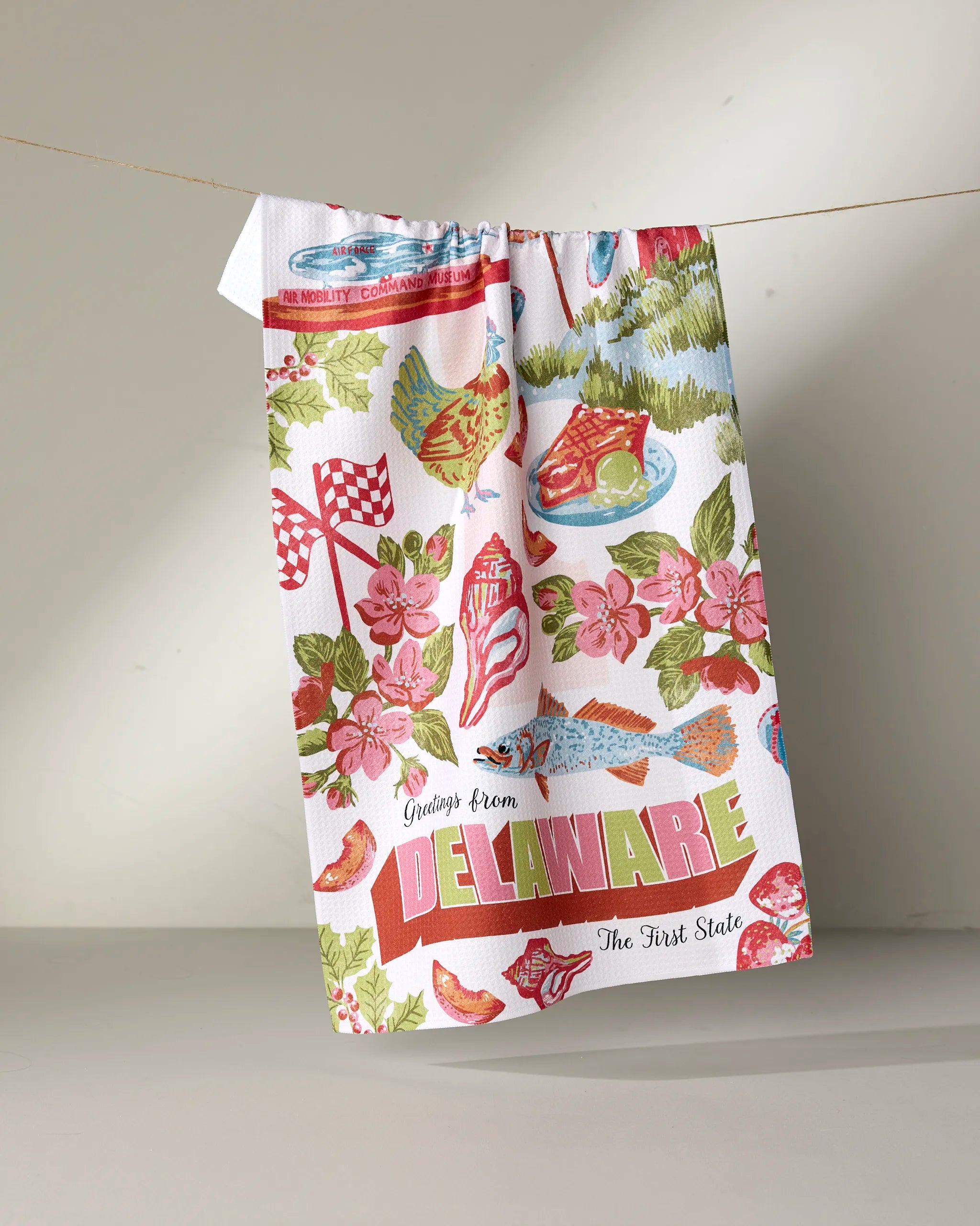 The Geometry “Greetings From Delaware” kitchen towel features vibrant illustrations of flowers, fish, and landmarks, plus bold “Greetings from DELAWARE The First State” text. Crafted from recycled materials for eco-friendly style.