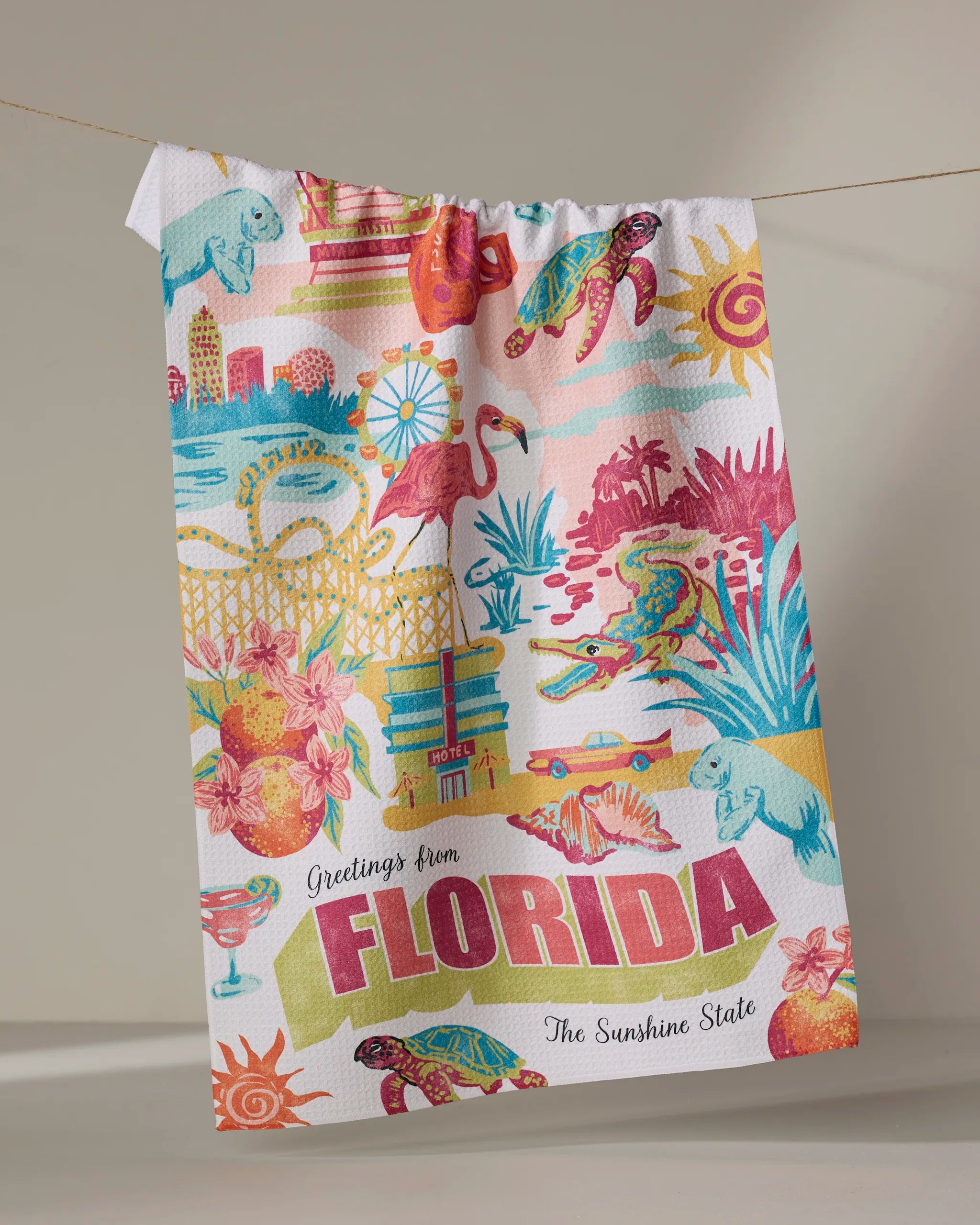 The Geometry "Greetings From Florida" tea towel, crafted from recycled materials, showcases vibrant Florida motifs—flamingos, turtles, citrus, beaches, hotels—and bold “Greetings from FLORIDA The Sunshine State” lettering.