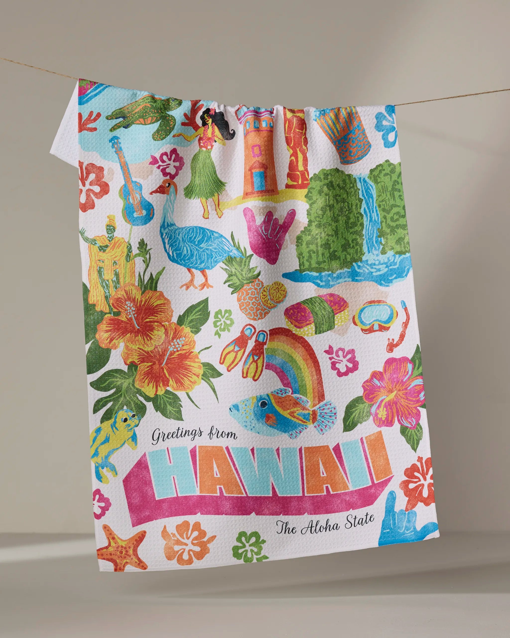 The Geometry "Greetings From Hawaii" tea towel features ultra-absorbent fabric and vibrant Hawaiian-themed designs—flowers, hula dancer, ukulele, and more—with "Greetings from HAWAII The Aloha State" text. Shown hanging on a line.