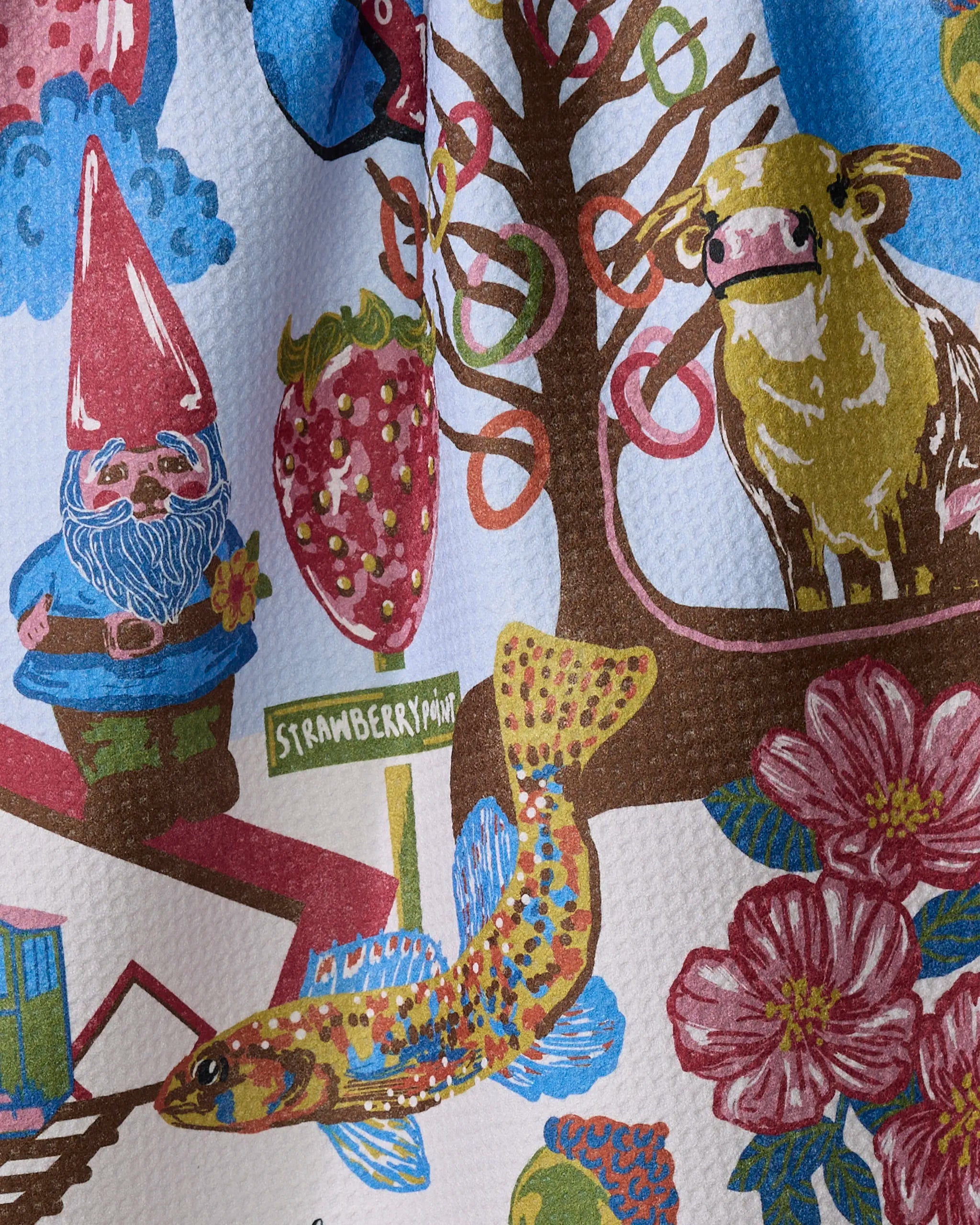 Geometry's "Greetings From Iowa" tea towel features a gnome, cow, strawberry, fish, flowers, tree, and a "Strawberry Point" street sign on a light background. Made from quick-drying recycled materials.