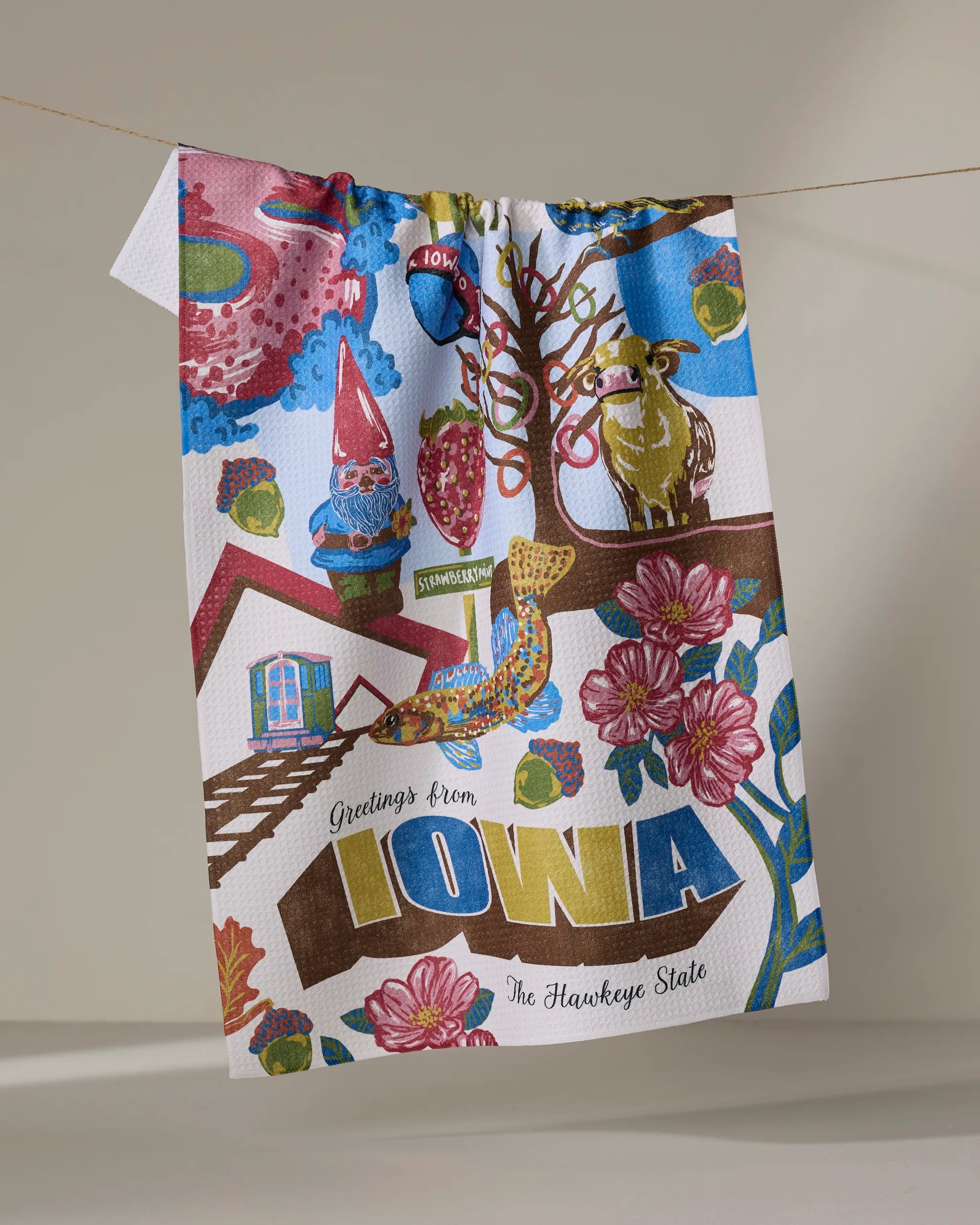The Geometry "Greetings From Iowa" tea towel dries quickly and features colorful illustrations and text like "Greetings From IOWA" and "The Hawkeye State," plus Iowa-themed images of flowers, barns, a cow, and a gnome.