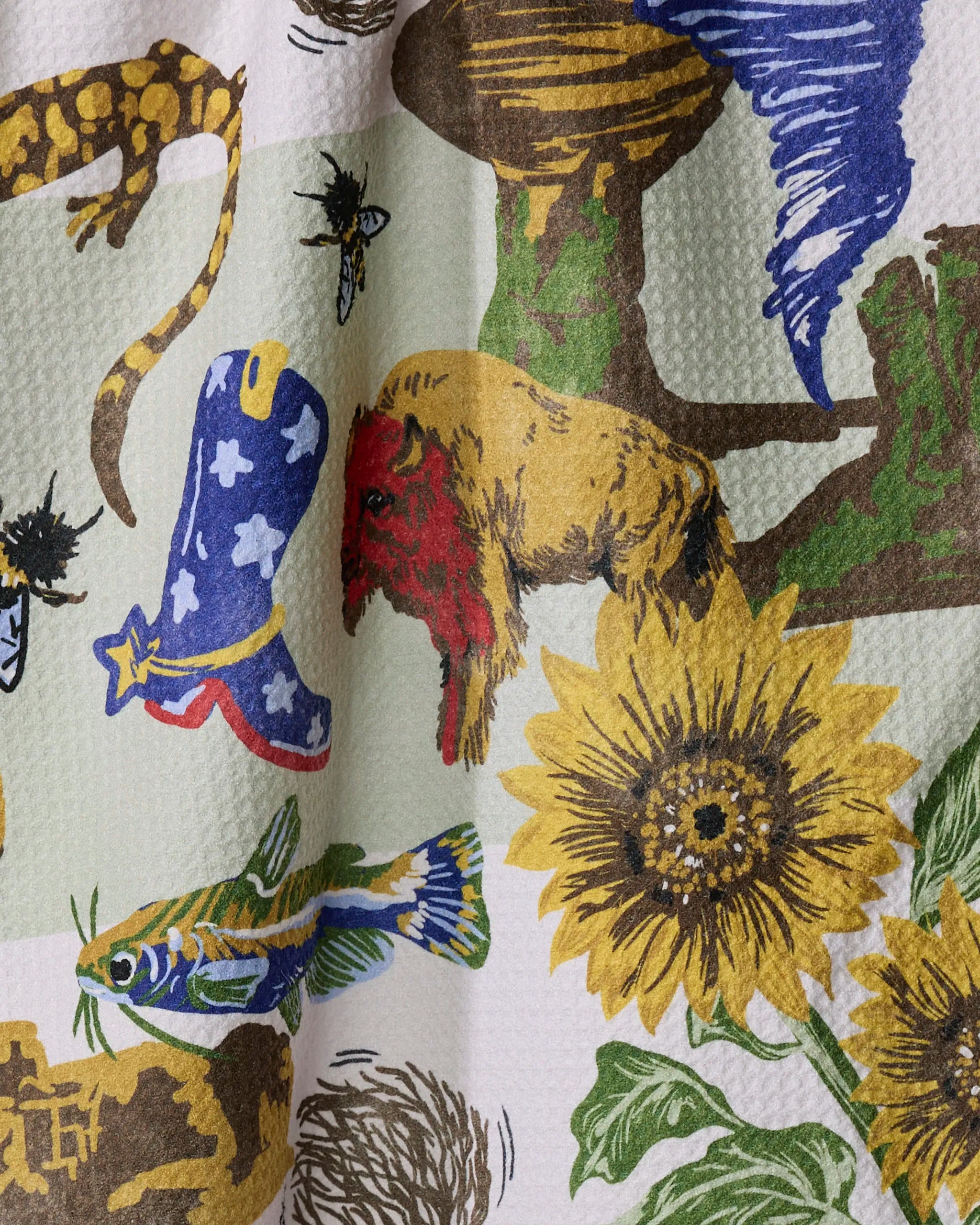 “Greetings From Kansas” by Geometry is a recycled kitchen towel with colorful illustrations—yellow sunflower, red and brown bison, blue cowboy boot, fish, insects, reptiles, and cacti—on a textured white background.