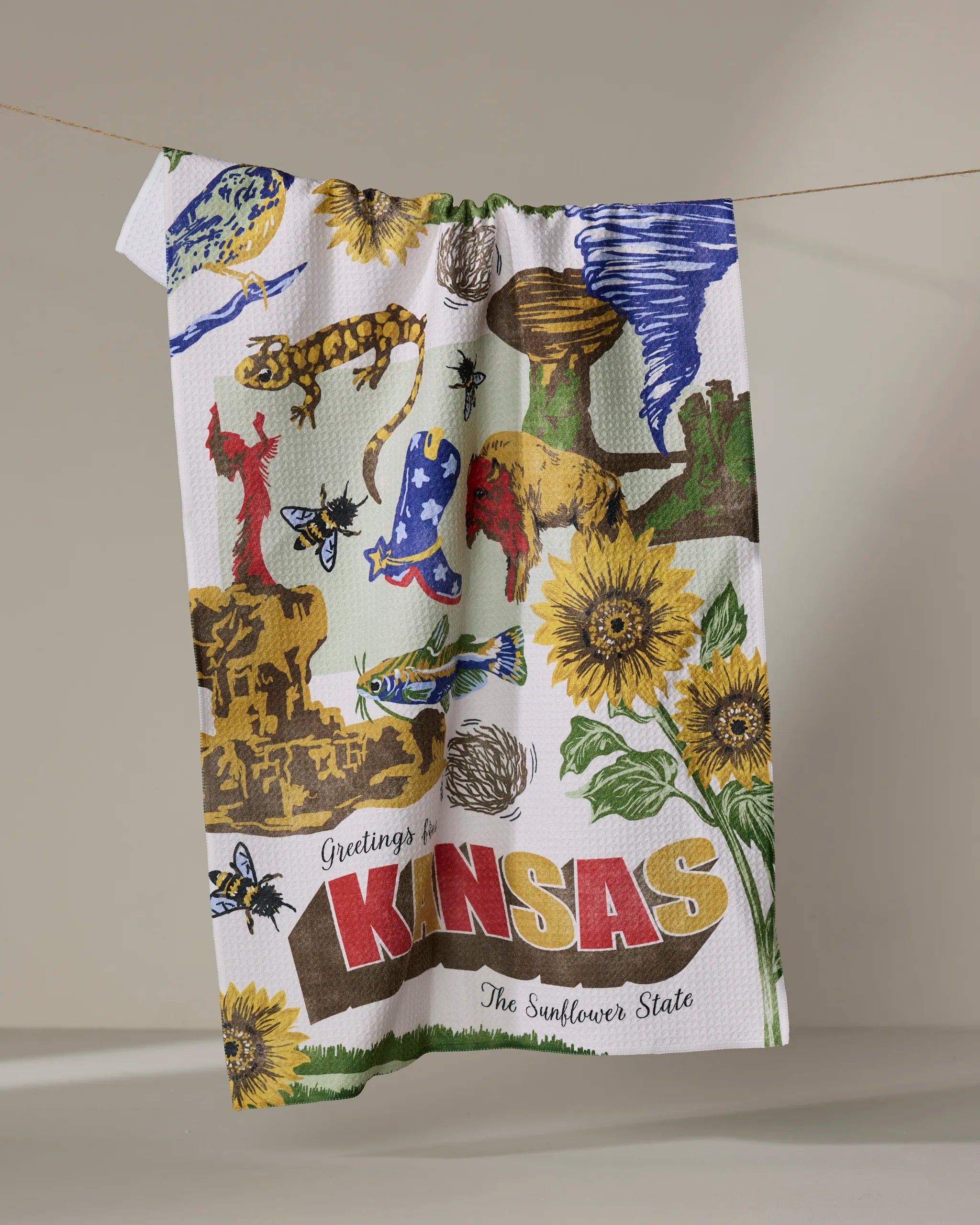 The Geometry "Greetings From Kansas" tea towel features colorful illustrations of Kansas landmarks, sunflowers, animals, and "Greetings from KANSAS The Sunflower State," adding charm and function to any kitchen. Ultra-absorbent design.