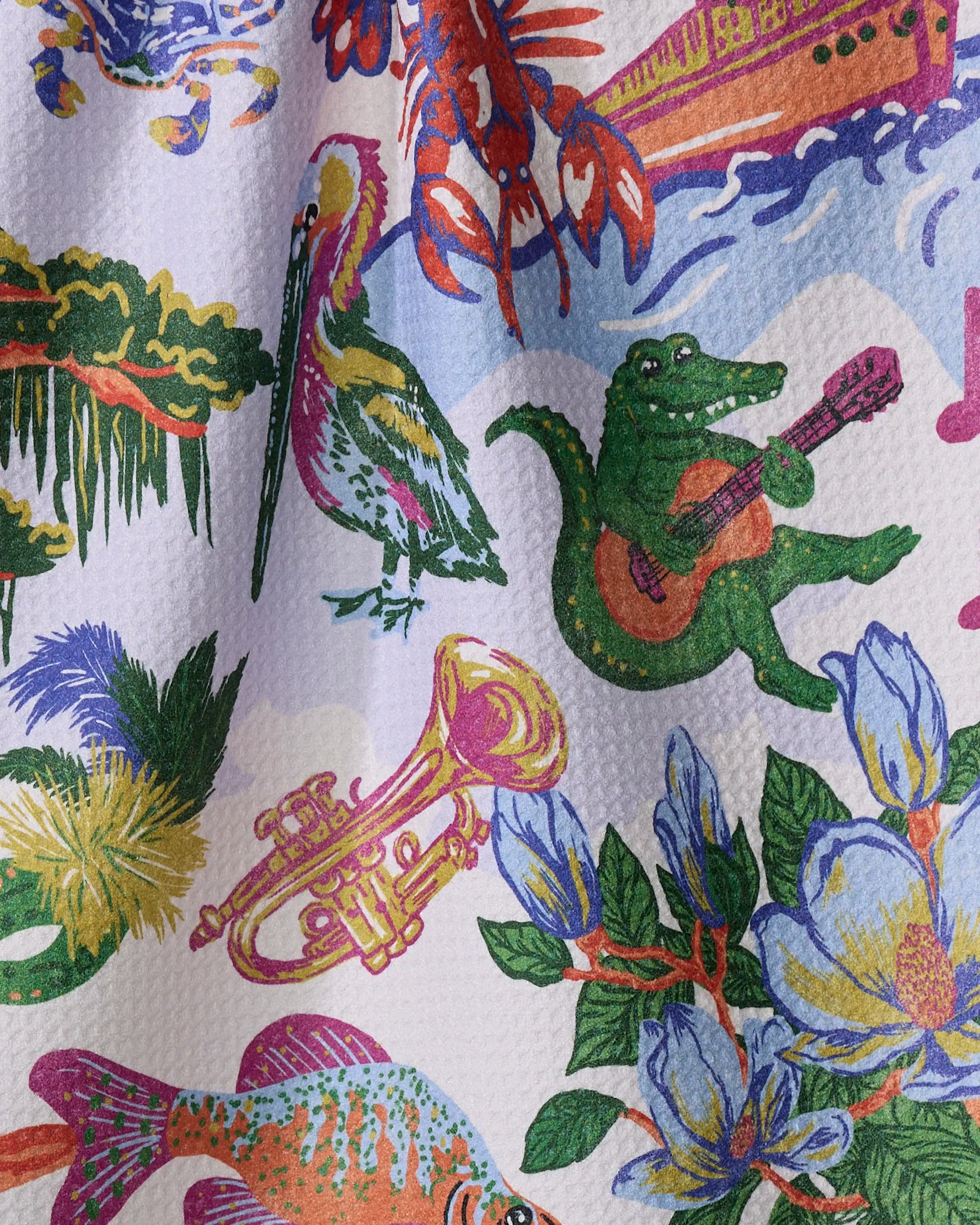 The "Greetings From Louisiana" recycled kitchen towel by Geometry features vibrant illustrations of a pelican, guitar-playing crocodile, lobster, fish, saxophone, magnolia flowers, and greenery on a textured white background.