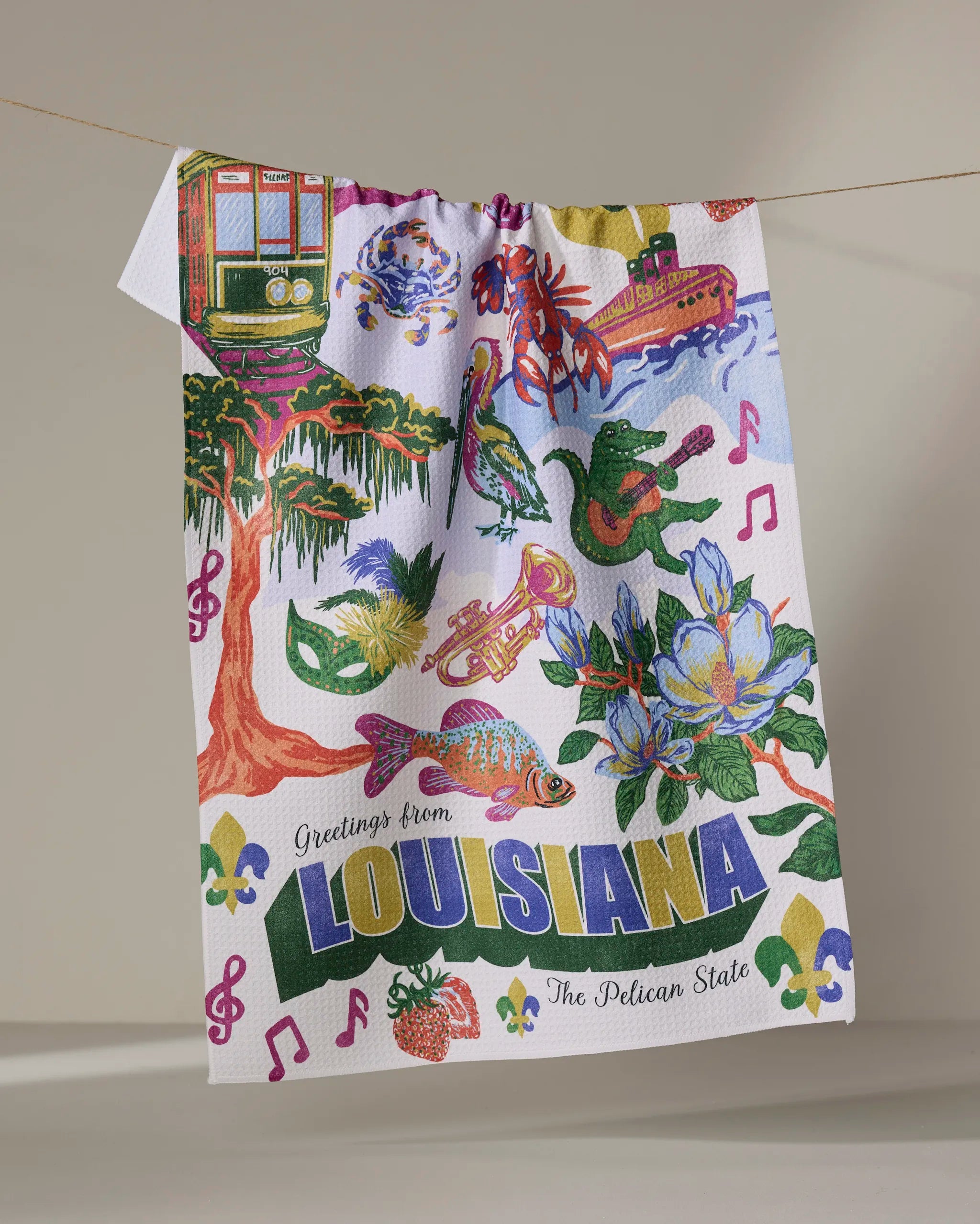 The Geometry Greetings From Louisiana tea towel features colorful illustrations—a streetcar, alligator, saxophone, crawfish, magnolia, riverboat, and musical notes—with the text "Greetings from Louisiana.