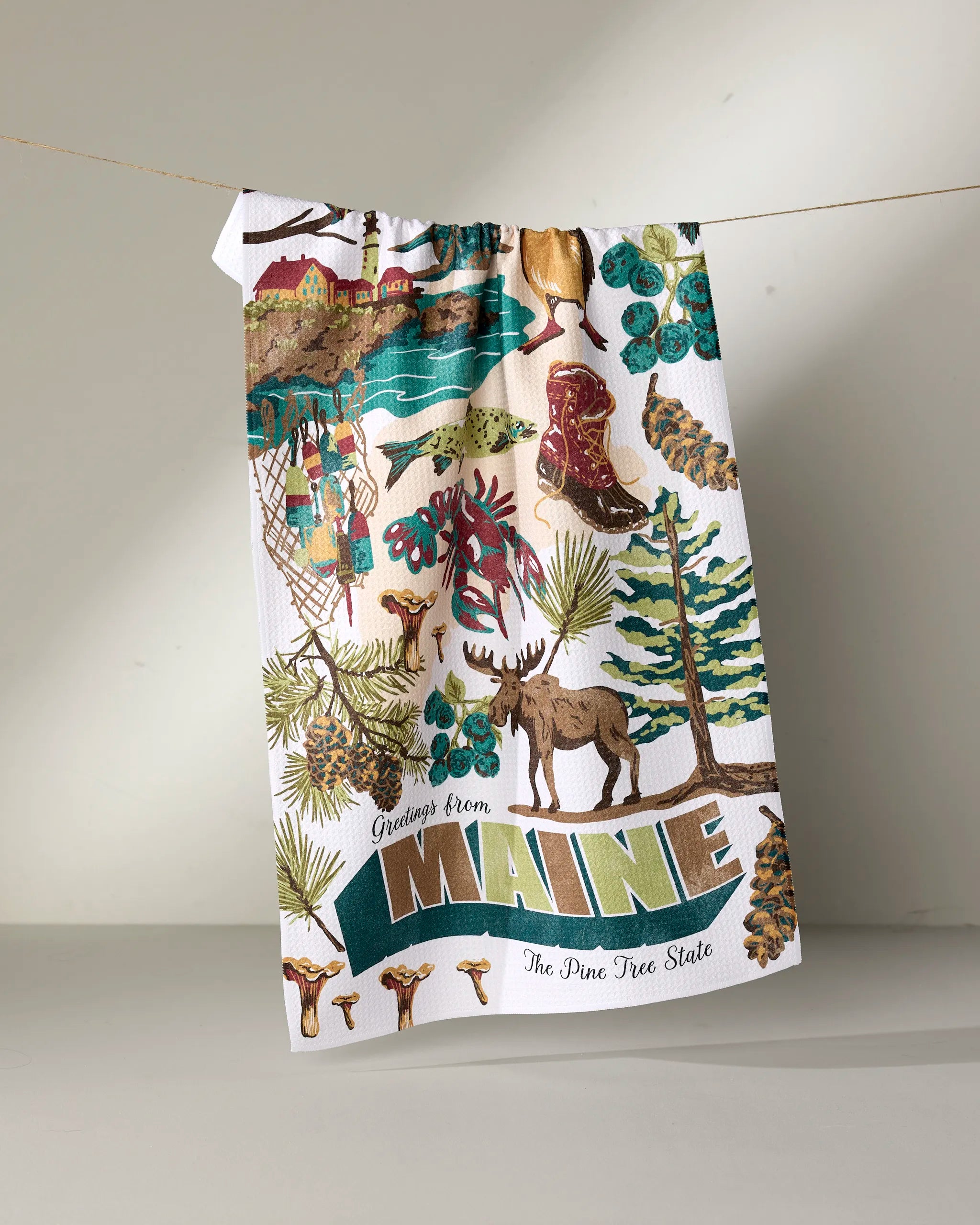 The Geometry “Greetings From Maine” tea towel displays vibrant images of a moose, pine trees, blueberries, a lighthouse, and forests with the phrase “The Pine Tree State,” made from recycled materials.