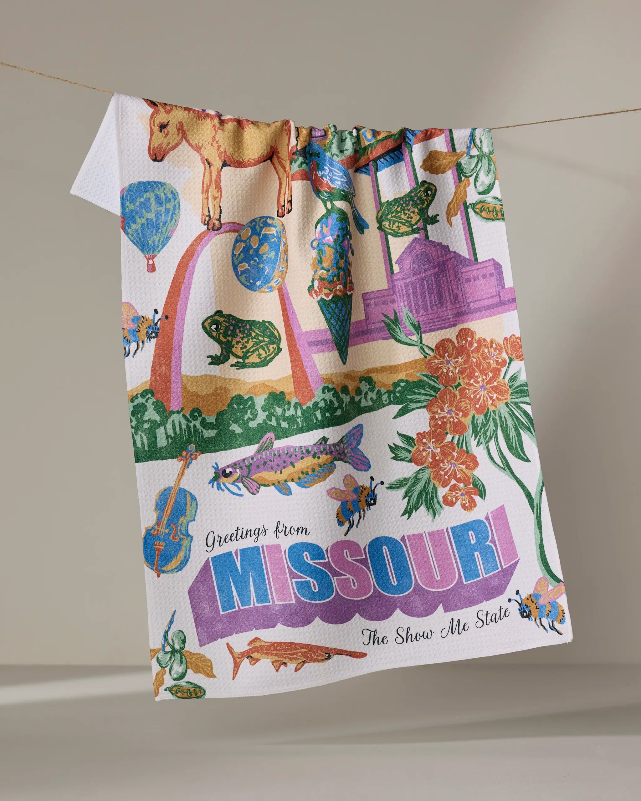 Geometry's "Greetings From Missouri" tea towel features vibrant illustrations of Missouri icons—flowers, instruments, salamander, hot air balloons, Gateway Arch—printed on soft Geoweave fabric with “Greetings from MISSOURI The Show Me State.”.