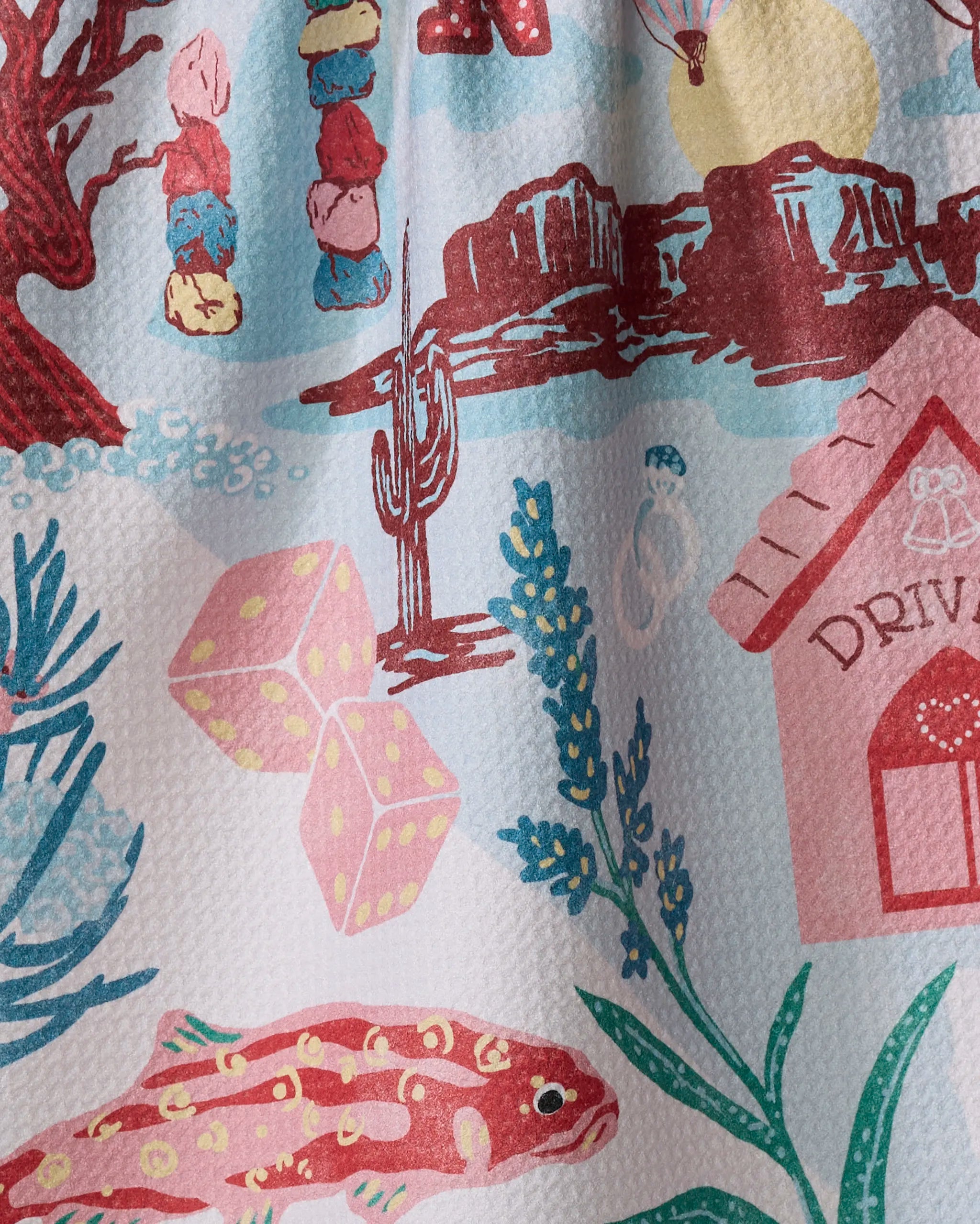 The "Greetings From Nevada" tea towel by Geometry showcases vibrant cacti, pink dice, a red fish, a drive-in cafe, red mountains, stacked rocks, and plants on a white background.