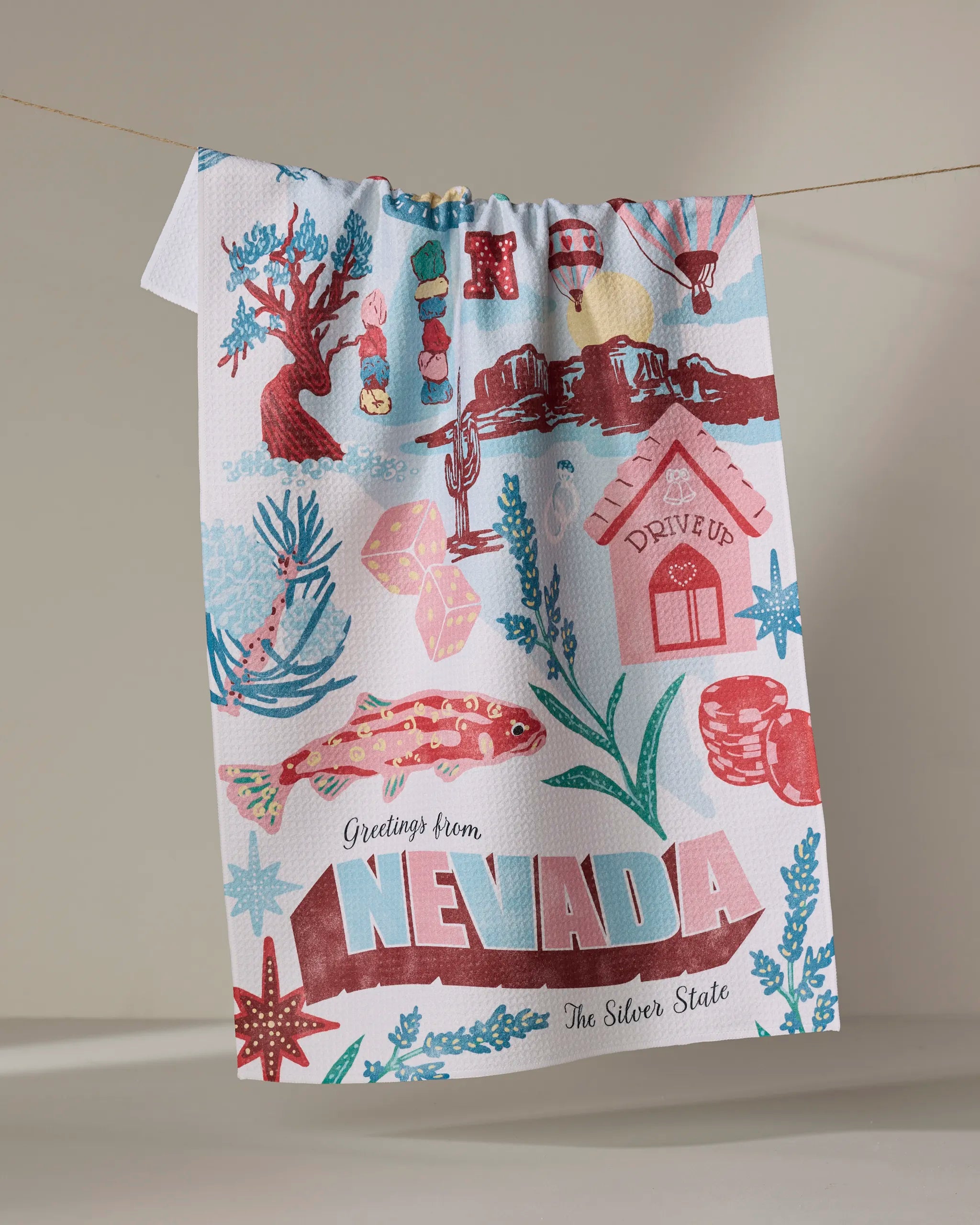 The Geometry Greetings From Nevada tea towel features colorful Nevada symbols—cactus, lizard, playing cards, gems, mountains, and bold “Greetings from NEVADA The Silver State”—and adds quick-drying charm to any kitchen.