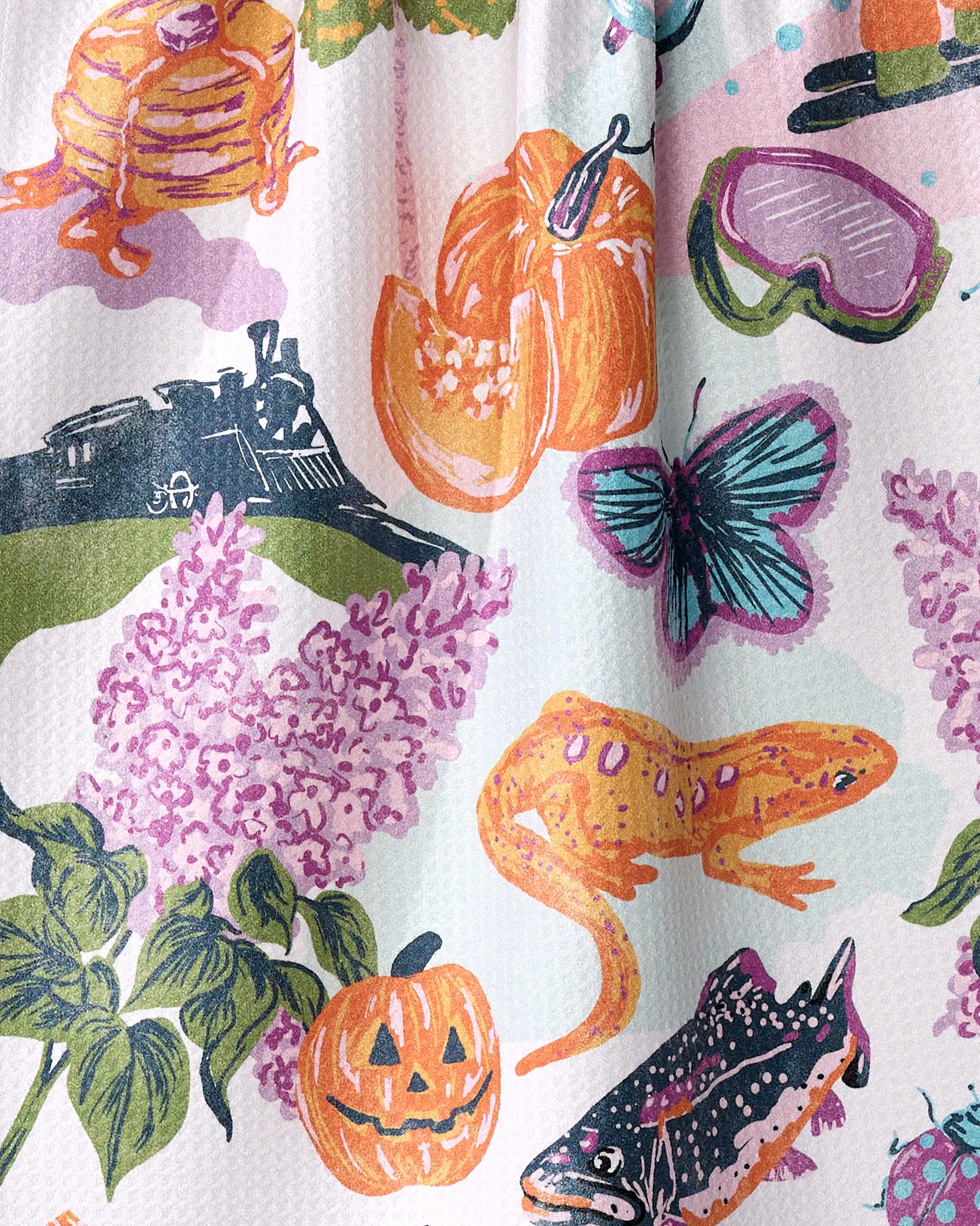 The Geometry "Greetings From New Hampshire" tea towel is colorful, quick-drying, made from recycled materials, and features illustrations of pumpkins, pancakes, a steam train, lilacs, butterflies, pie, a salamander, fish, jack-o’-lanterns & swim goggles.