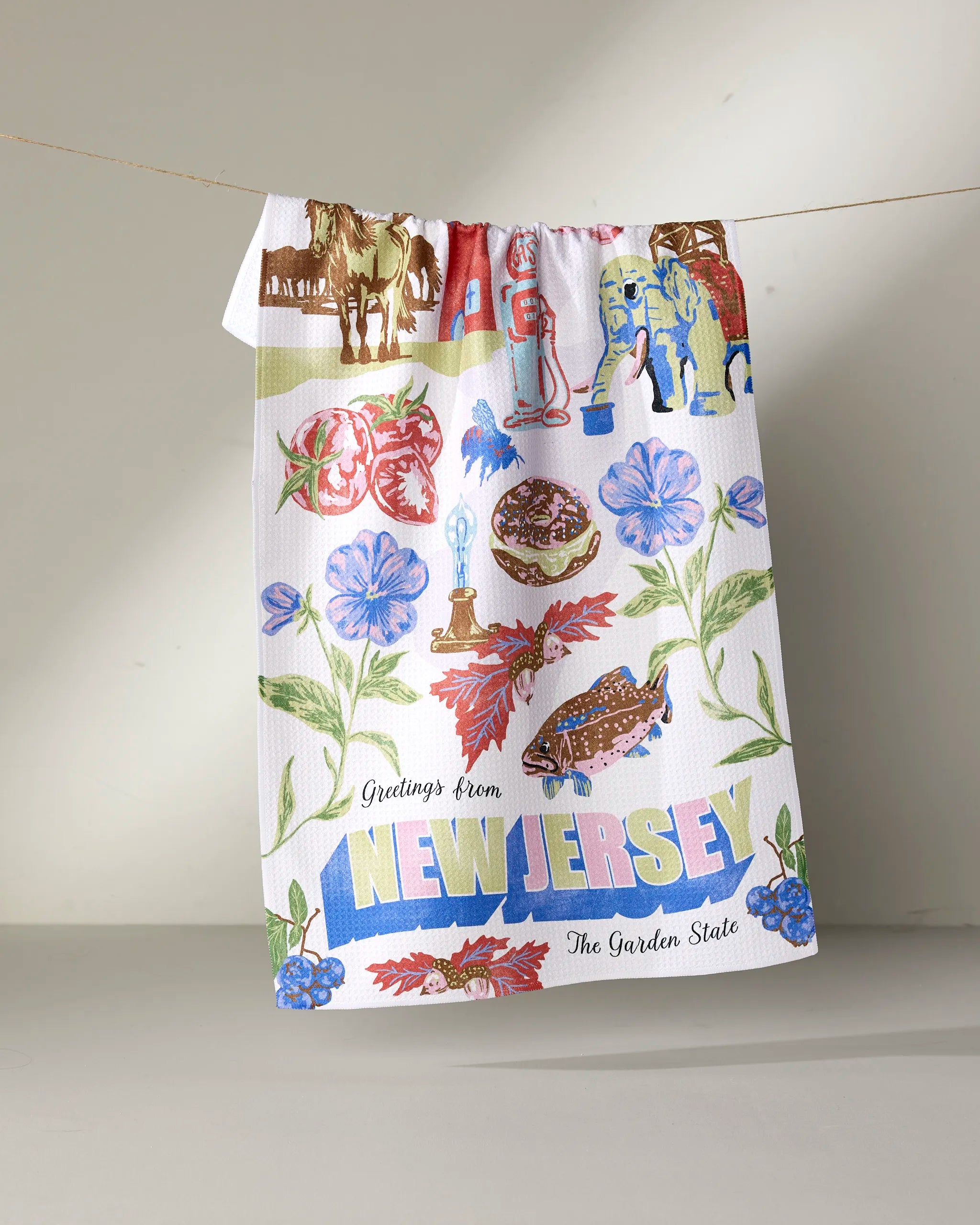 The Geometry "Greetings From New Jersey" tea towel, made from ultra-absorbent recycled materials, features colorful state-themed illustrations and the phrase "Greetings From NEW JERSEY The Garden State.