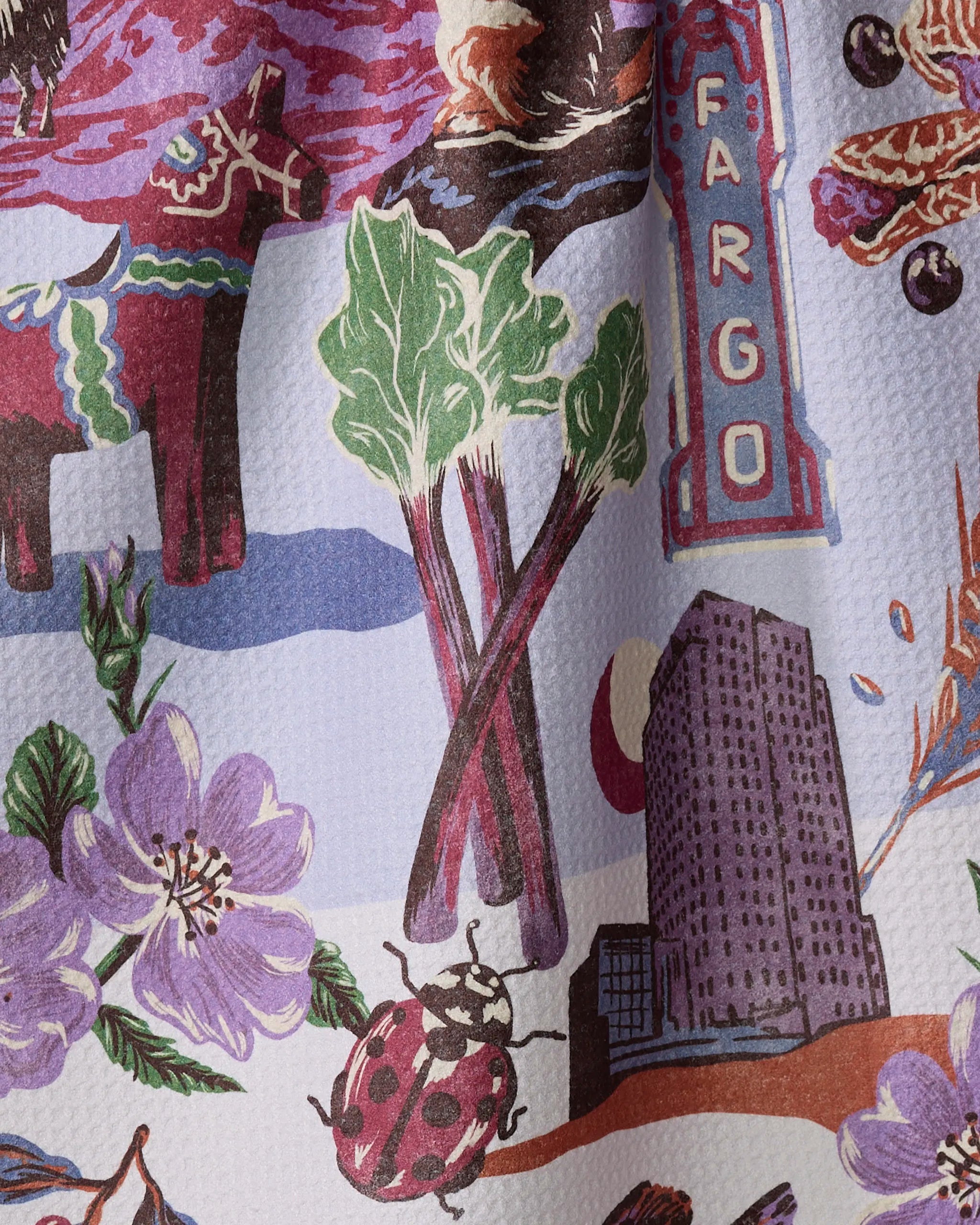 The Geometry "Greetings From North Dakota" dish towel features an illustrated pattern with rhubarb stalks, a Fargo sign, a tall building, a ladybug, purple flowers, a colorful cow, and other local motifs on a textured background.