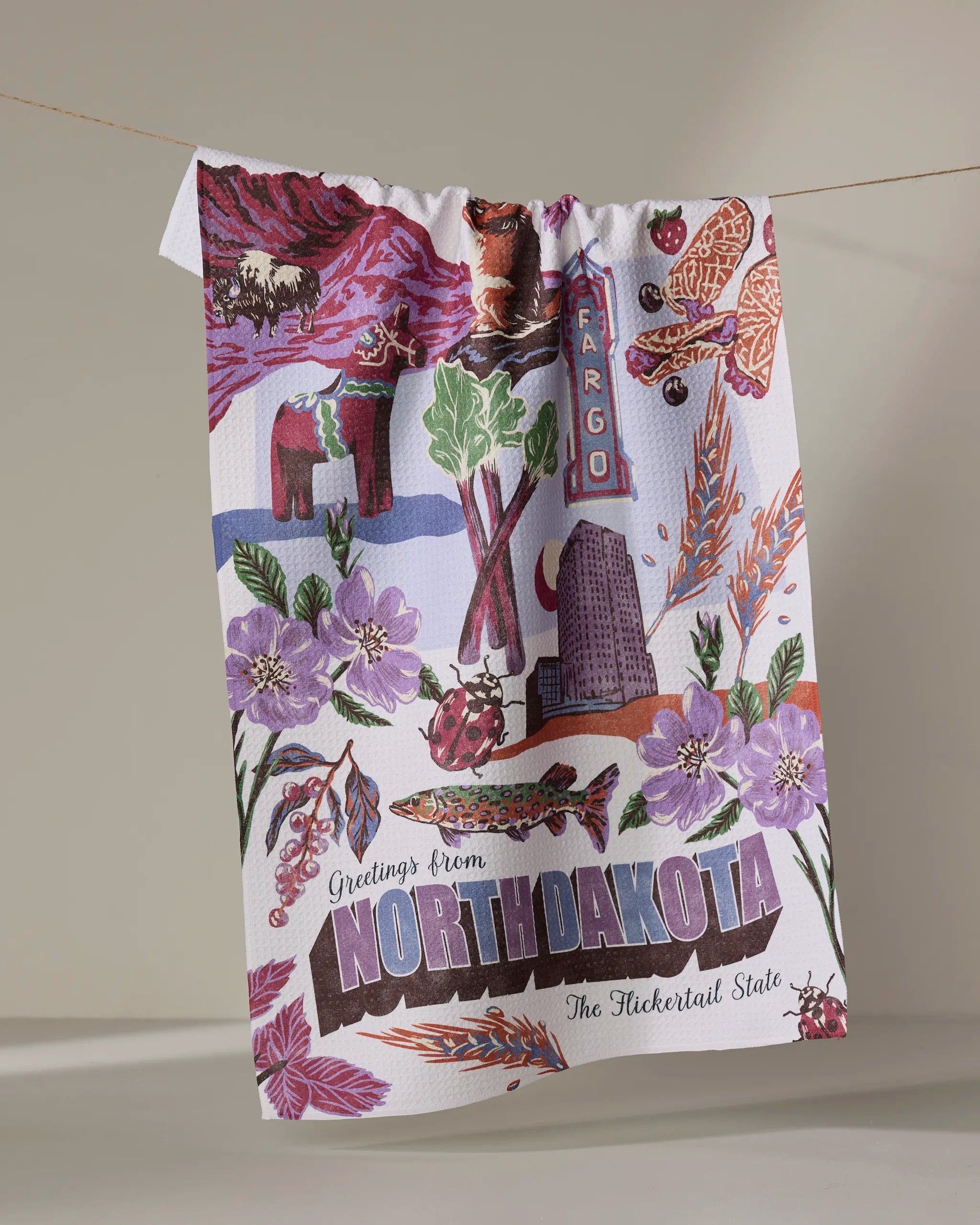 A vibrant Geometry "Greetings From North Dakota" recycled kitchen towel hangs on a line, showcasing deer, bison, fish, flowers, grain, landmarks, and the text "Greetings From North Dakota: The Flickertail State.